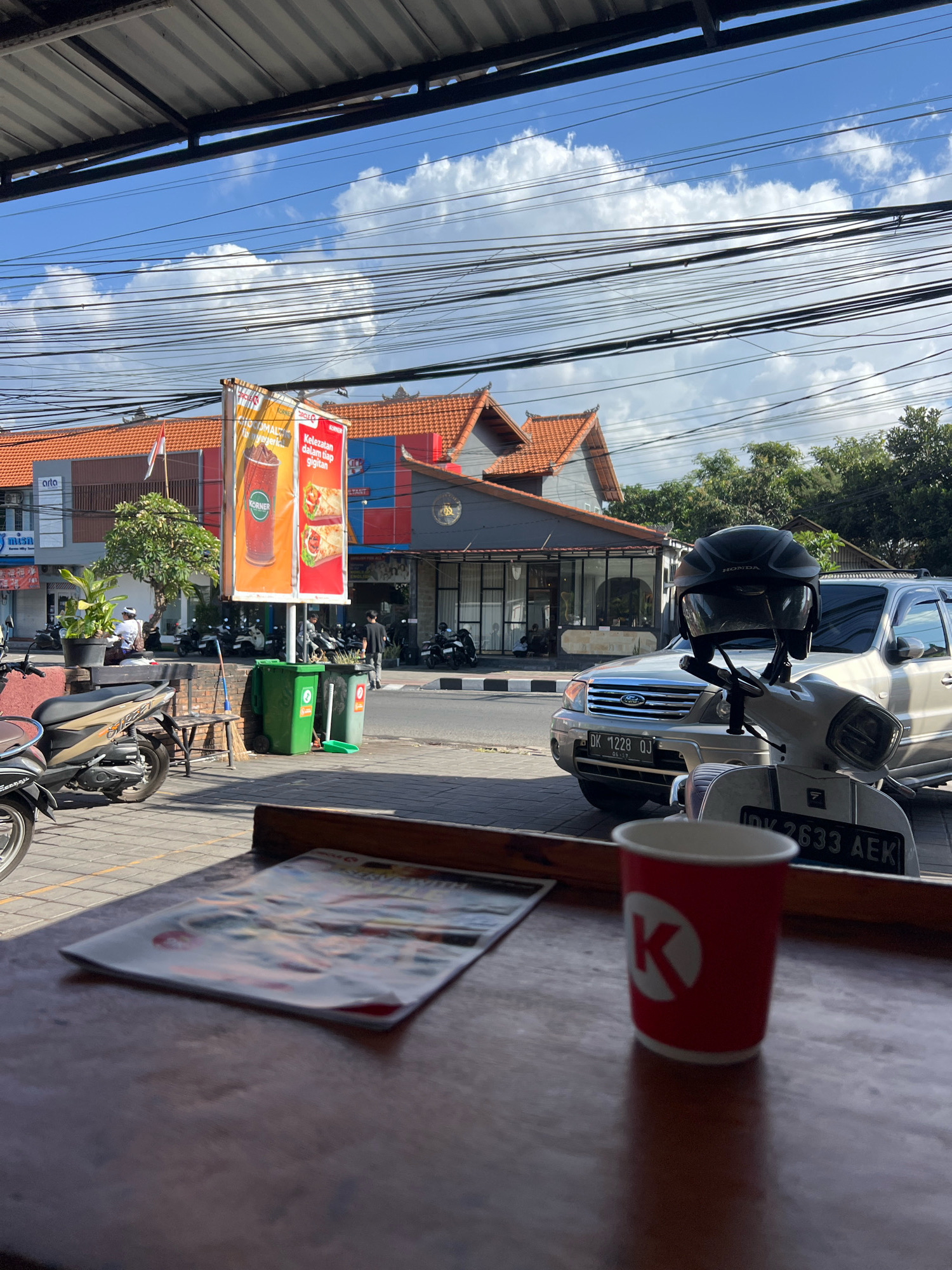 The image depicts a street scene in Denpasar, Indonesia, on a bright, sunny day with scattered clouds. The foreground features a wooden table with a red paper cup with a white "K" logo on it and a magazine or pamphlet. Behind the table, a sidewalk is visible, paved with interlocking bricks. To the left, a portion of a beige and brown scooter is parked.
Beyond the sidewalk, a street runs horizontally. On the other side of the street, there are several buildings with orange tiled roofs and grey facades. One building has a large advertisement poster displaying images of drinks and food. Several motorcycles are parked in front of the buildings.
A light-colored SUV with license plate "DK 1228 QJ" is parked on the street. A white scooter with a black helmet on its handlebars and license plate "DK 2633 AEK" is parked in front of the SUV. Green and grey trash bins are situated on the sidewalk near the advertisement poster. A broom and dustpan are placed next to the trash bins.
In the background, lush green trees and dense power lines crisscross the sky, indicating an urban environment. The overall impression is a bustling street scene, possibly near a cafe or shop, with a mix of transportation and commercial activity. There are a few individuals visible in the mid-ground, some walking and others seated, but their actions are not clearly discernible. The sunlight casts shadows, indicating the time of day is likely mid-morning or afternoon.