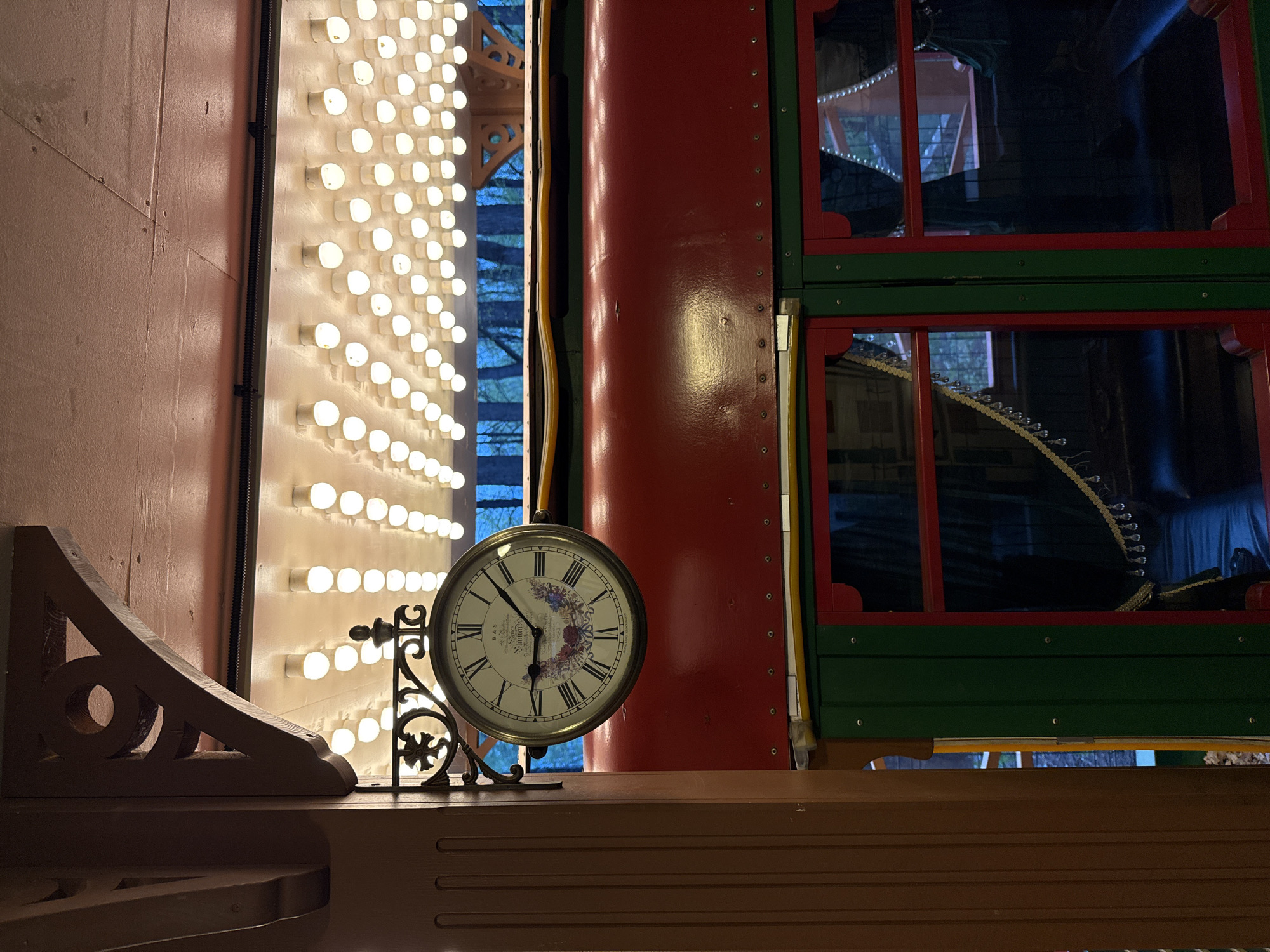 The image shows an indoor scene featuring a decorative clock in the foreground. The clock has a white face with Roman numerals and a floral design in the center. It is mounted on an ornate, dark-colored metal stand. To the left of the clock is a wooden corbel or bracket with a circular opening.
Behind the clock and to the left is a wall adorned with rows of illuminated light bulbs, creating a bright, theatrical effect. To the right of the clock is a structure with red and green framing, possibly a window or decorative element, that appears to be part of a larger architectural feature or display. The colors suggest a festive or carnival-like atmosphere.
The objects are resting on a wooden surface, possibly a shelf or counter. The lighting is a mix of natural light from the window or door and the artificial light from the bulbs, creating a contrast in brightness and shadows.
Without additional visual context, the precise location within Perm, Russia, cannot be definitively identified, but the image suggests a theatrical, recreational, or commercial setting based on the presence of the marquee lights and colorful architectural elements.