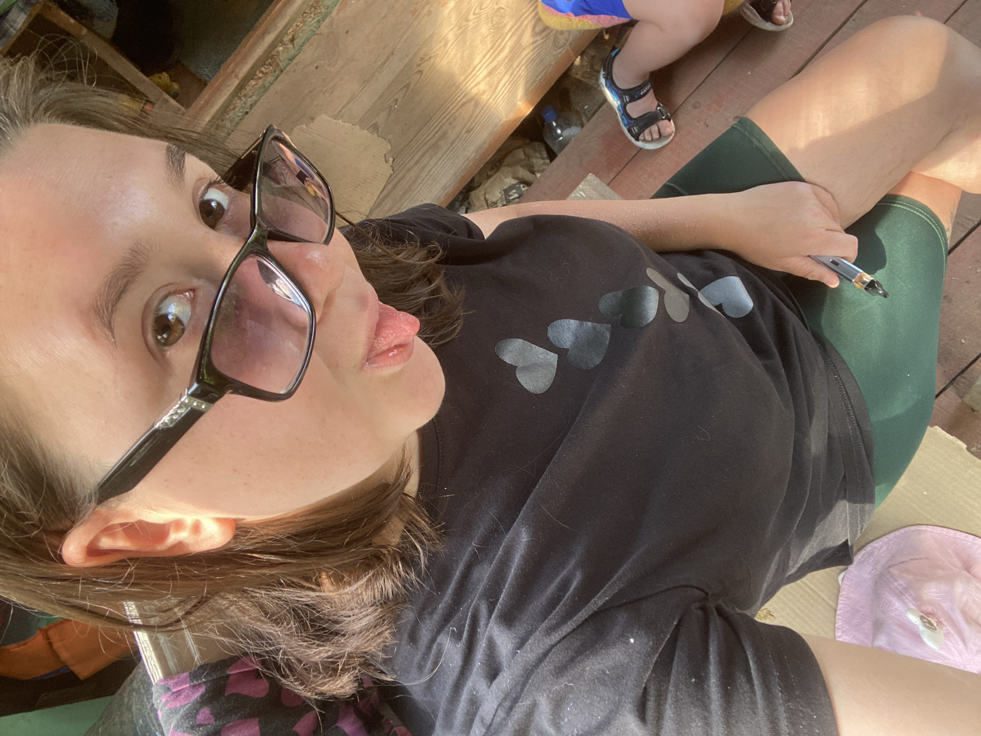 The media file is a close-up, over-the-shoulder selfie of a woman. She is wearing large black-rimmed glasses and a black t-shirt with a heart design. Her tongue is sticking out, and she appears to be playful or making a silly face. In the background, there are hints of a casual, outdoor setting, possibly a porch or deck with wooden planks. A child's feet wearing sandals and part of their colorful clothing are visible in the upper right. A plastic water bottle and what appears to be a vape pen are also present in the scene. The lighting suggests it is daytime. The overall mood is lighthearted and informal. The woman is holding a silver and black vape pen in her right hand, which rests on her knee. Her legs are crossed, and she is wearing dark green shorts. A pink hat is lying on the ground to her right.
