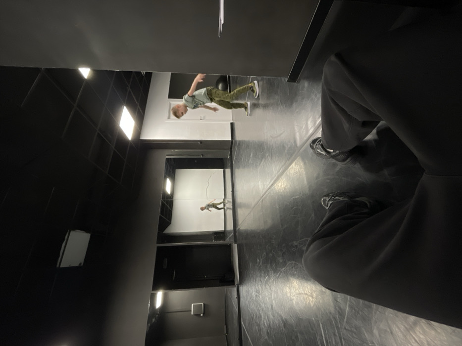 A person is reflected in a mirror with white walls and bright lights. The image is taken from a high angle, showing a portion of someone's lap, shoes, and legs in the foreground. The walls and floors of the room are a mix of black and white, with geometric patterns on the ceiling. The overall scene appears to be a dance studio or theater space.