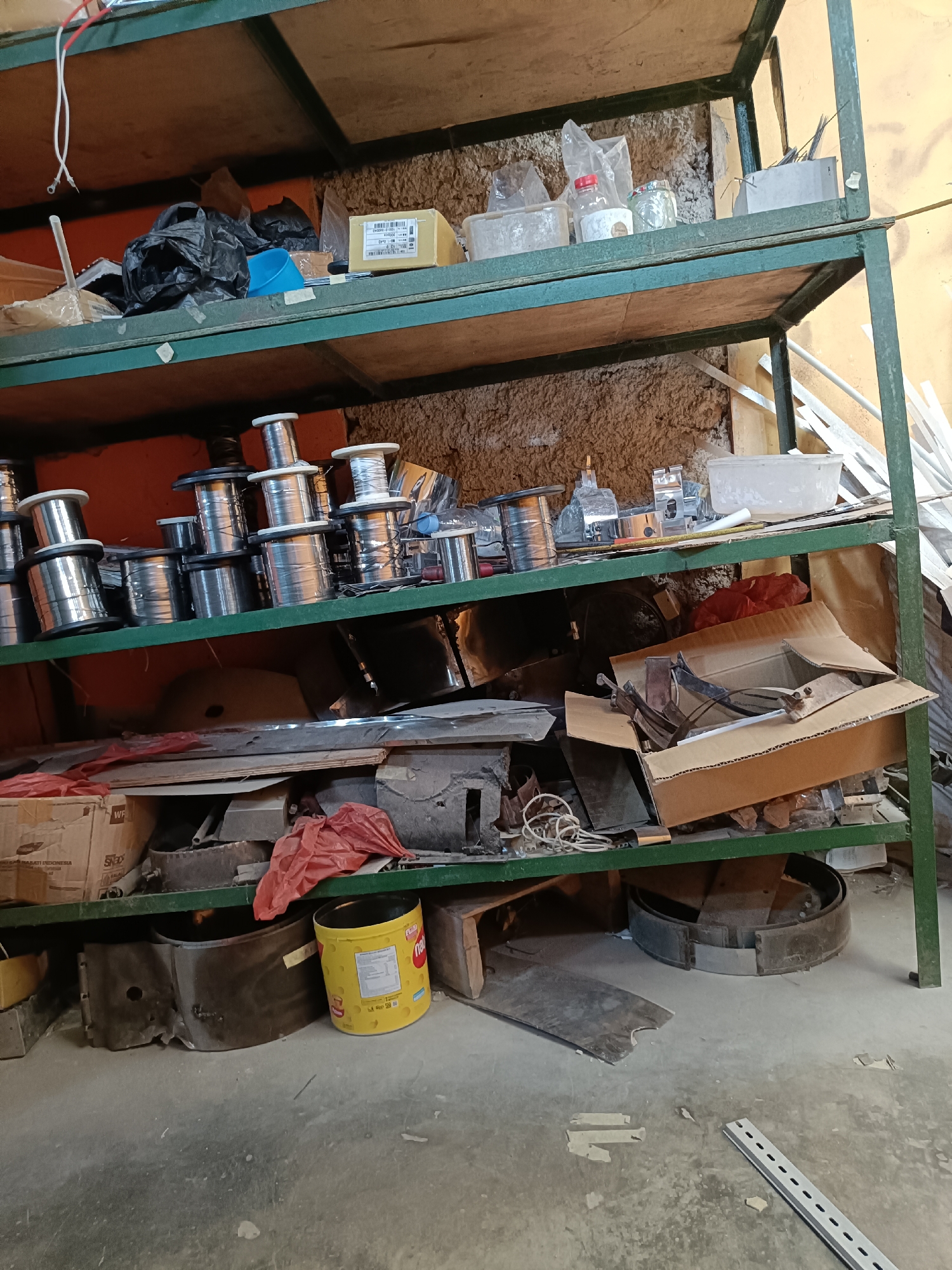 The image depicts a cluttered storage area, possibly a workshop or a shed, located in Telagamurni, Indonesia. The scene is dominated by a multi-tiered metal shelving unit filled with various items.
On the top shelf, there are several black plastic bags, a light blue plastic cup, and some cardboard boxes, one of which appears to be labeled with text that includes "INDONESIA". A white and red wire hangs down from the left side of this shelf.
The second shelf from the top contains numerous spools of wire, stacked both horizontally and vertically. These spools are primarily made of metal, with some appearing to be silver or chrome. In addition to the wire spools, there are some plastic containers, glass jars, and what appear to be metal components or tools.
The third shelf is more varied. It holds more wire spools, some smaller items, and a larger, open cardboard box containing metal parts and possibly tools. A red plastic bag is also present on this shelf.
The bottom of the shelving unit and the floor below are also visible. There are large metal rings or brake drums, a yellow container with labels, and various pieces of metal and debris scattered on the floor. The wall behind the shelves has a rough, textured finish. The overall impression is one of a functional but disorganized storage space.