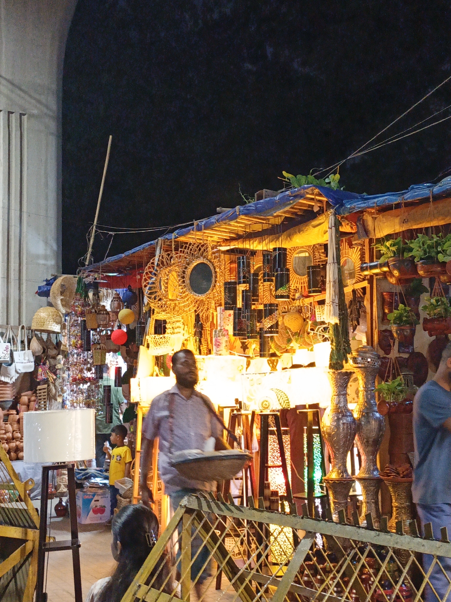 An outdoor market stall at night is illuminated by various lamps and string lights, displaying a variety of handicrafts, including woven baskets, decorative mirrors, hanging lanterns, and vases. A man, possibly a vendor, stands in the foreground holding a tray, while a young boy in a yellow shirt looks on from behind a white lampshade. A woman is partially visible in the lower left corner, facing away from the camera. The stall is covered with a blue tarp, and the backdrop is a dark, starry sky.