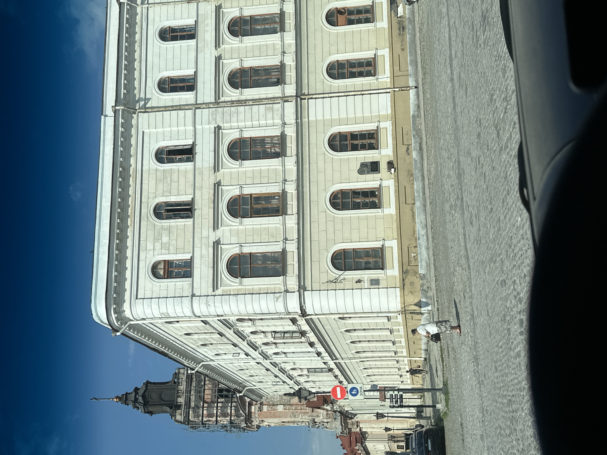 The image shows a broad view of a street in Kamianets-Podilskyi, Ukraine, featuring a large, pale yellow building with multiple arched windows and intricate architectural detailing, possibly a historical landmark or administrative building. A tall, dark tower with a copper spire, partially covered in scaffolding, rises to the left of the main building, suggesting ongoing construction or renovation. The street is paved with cobblestones, and several lampposts are visible. In the foreground, a woman wearing a white shirt and a patterned skirt stands on the street, holding a dark handbag, and appears to be looking away from the camera. A black car is parked on the left side of the street. The sky is a clear, deep blue.