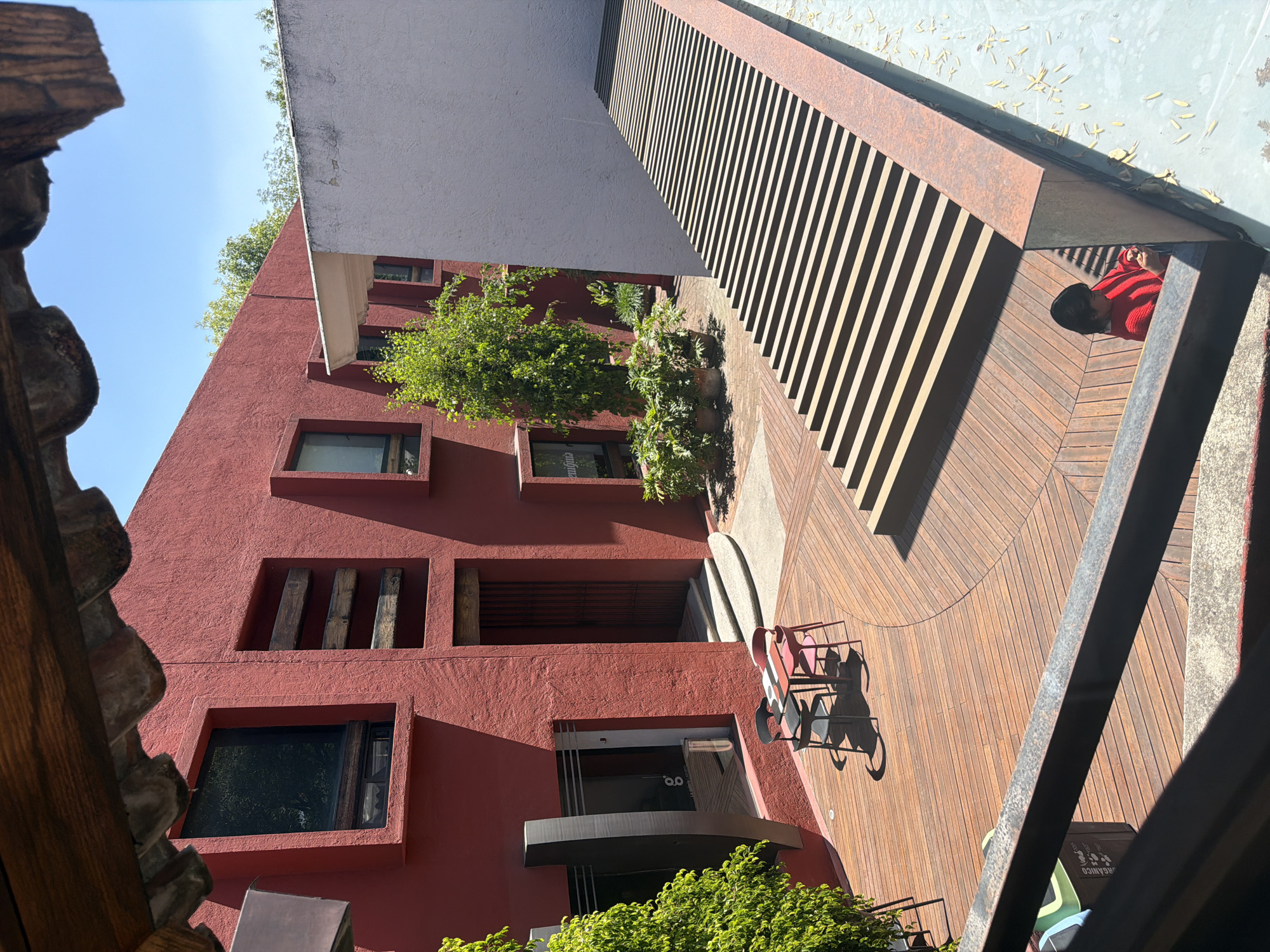 The image depicts an elevated view of an open-air courtyard and building complex identified as La Galería La Ceiba in Puebla, Mexico. The primary structure features multi-story, textured red stucco walls, punctuated by rectangular dark-framed windows and recessed rectangular niches, some containing horizontal wooden beams. Signage reading "la galería La Ceiba" and "enigma" is visible on ground-level windows. A main entrance with a dark metal gate is situated at the base of the red building, accessible via curved concrete steps.
The courtyard floor is predominantly composed of wooden decking laid in a circular pattern, with a smaller paved section deeper within the complex. On the wooden decking, a square outdoor table with a black and red patterned top is surrounded by four chairs: two black and two pink. A dark grey rectangular waste bin, partially visible on the left, is labeled "ORGANICO".
To the right, a tiered slatted wooden structure extends alongside a plain light grey textured wall. Below this structure, in the lower right, a shallow, light blue water feature contains scattered yellow leaves. A female individual, partially obscured by a dark metal railing, is positioned beside this water feature. She has dark hair, wears a red long-sleeved top, and is observed looking at a small handheld device.
Lush green foliage, including a tall tree on the left and various potted plants, is distributed throughout the courtyard. The scene is illuminated by direct sunlight under a clear blue sky, with dark wooden eaves framing the top of the image.