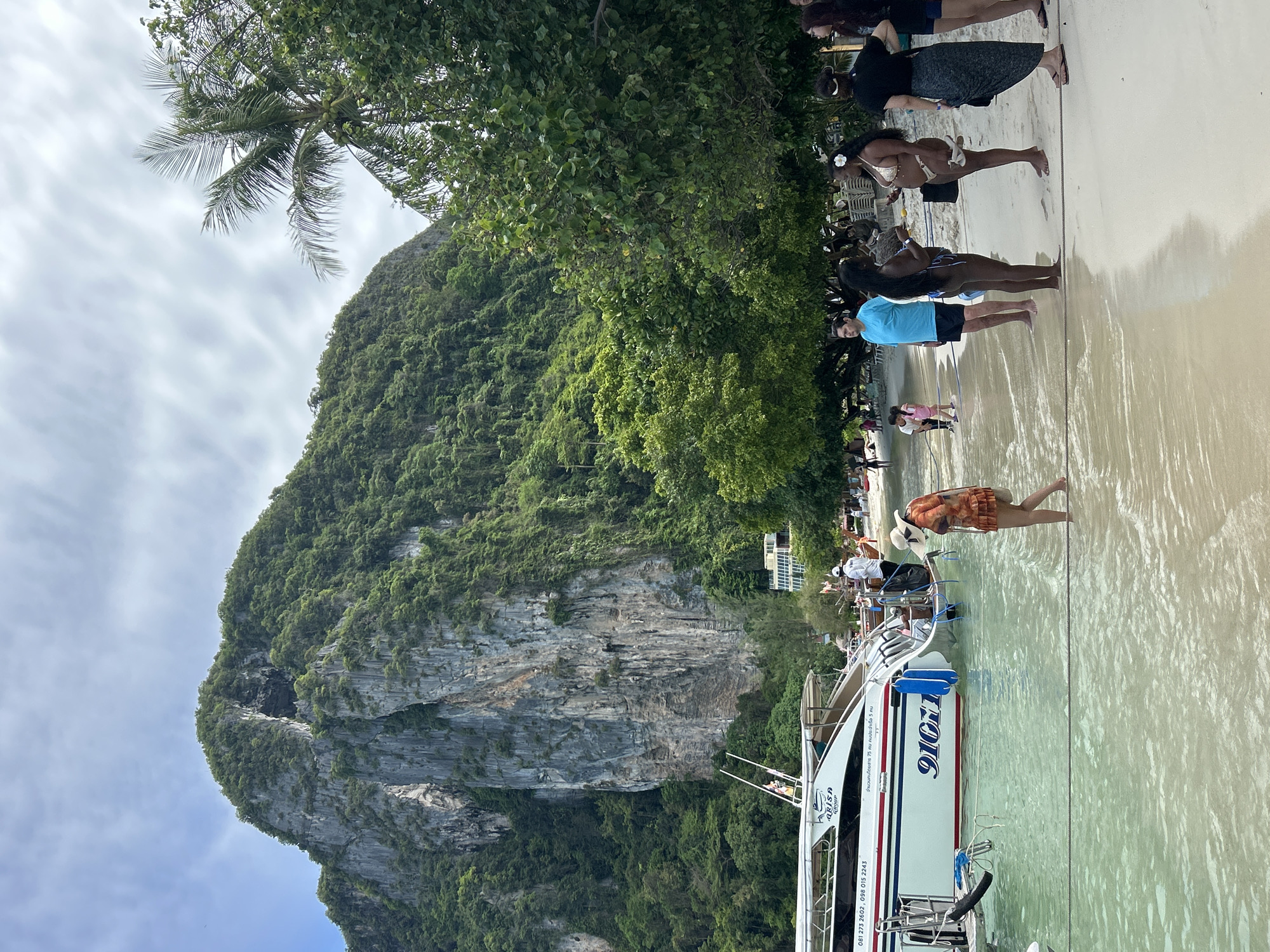 The image depicts a crowded tropical beach scene in Ao Nang, Thailand, characterized by towering limestone karsts and clear turquoise water. The upper portion of the frame is dominated by a cloudy sky with patches of light grey-white and faint blue. A colossal, steep limestone karst, densely covered in vibrant green tropical vegetation, occupies the left and central background. This karst features prominent vertical grey rock faces and a large, dark, irregular cave entrance near its summit. To the right, another section of the karst is visible, partially obscured by trees. The mid-ground features a line of lush tropical trees, including a palm tree on the right laden with coconuts, bordering the sandy beach. Nestled at the base of the karst, to the right of the main boat, a modern, multi-story building with light-colored walls and dark windows is visible, suggesting a resort or guesthouse. The lower portion of the image shows a light-colored, sandy beach with shallow, clear, turquoise-green water lapping at the shore. The water exhibits ripples and slight turbidity from suspended sand. A thin black rope is stretched horizontally across the lower frame, partially submerged in the water, separating the immediate foreground from where people are standing. Multiple people are present on the beach and in the water. On the left, a large, white speedboat, marked "ARISA GROUP" and bearing the numbers "91011" in blue, is moored in the shallow water. Its hull also displays Thai text indicating a passenger capacity of 75 people and a crew of 5, along with two phone numbers (081 273 2602, 098 015 2243). A woman wearing a wide-brimmed straw hat and an orange patterned cover-up wades barefoot in the water near the speedboat, facing left. Further down the beach to the left, a small child in a pink outfit walks on the sand, and indistinct figures occupy beach chairs under trees. On the right side of the frame, a group of five adults stands barefoot on the wet sand near the waterline. A woman with dark hair, wearing a light-colored two-piece swimsuit and a white flower in her hair, holds white sandals and engages in conversation with another woman, also in a two-piece swimsuit. A man in a blue t-shirt and dark shorts stands to their left, looking towards the left side of the frame. Another woman with dark hair tied back wears a black t-shirt and a dark patterned