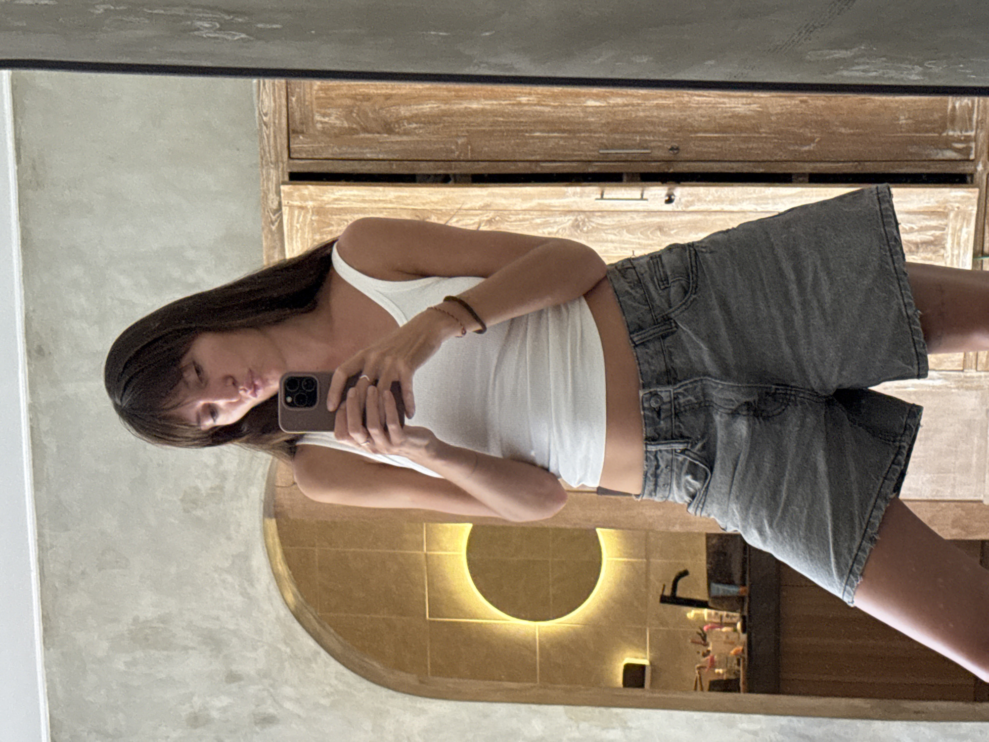 In North Kuta, Indonesia, a young woman with long, dark hair and bangs is captured in a mirror selfie taken in a bathroom. She is wearing a white tank top and grey denim shorts. Her left leg is slightly extended forward. She holds a phone in her right hand, obscuring part of her face as she pouts her lips. The background reveals a modern bathroom with concrete walls, an arched alcove with warm lighting, and a wooden cabinet.