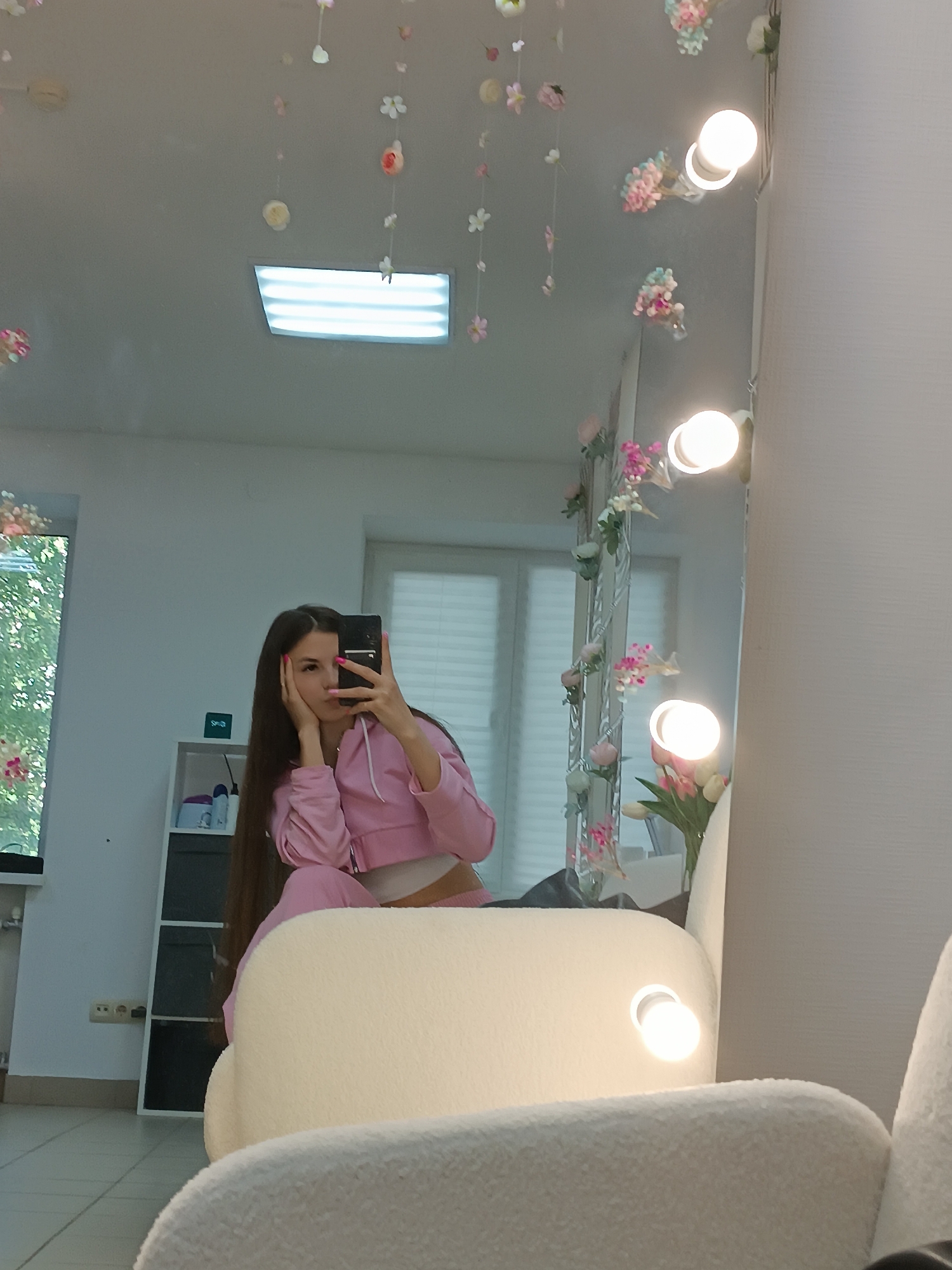 The image shows a person taking a selfie in front of a mirror decorated with lights and artificial flowers. The person, likely a young woman with long, dark hair, is wearing a pink tracksuit. She is holding a smartphone, partially obscuring her face. The mirror's reflection shows a room with a window, a cabinet, and another window with blinds. The room is well-lit, with additional lighting coming from a rectangular ceiling light. A white, textured armchair is in the foreground. The location is indoors, potentially a dressing room or a personal space. The specific city and state are unknown.