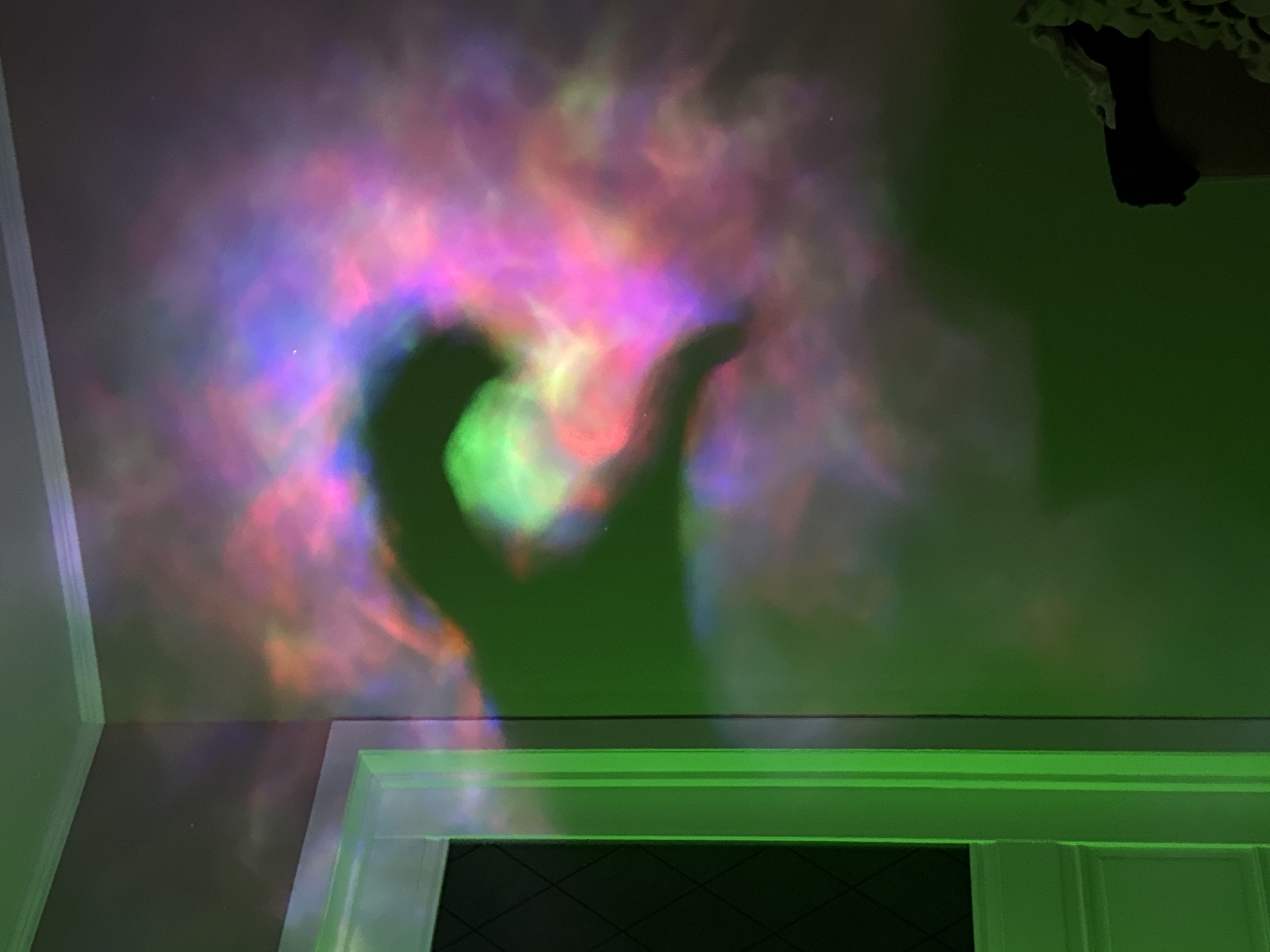 A low-angle shot depicts a wall with multicolored nebulous light projections predominantly in shades of pink, purple, and green. A dark, silhouetted figure stands in front of the light, likely casting a shadow on the wall. The lower portion of the wall is defined by a green-lit doorway or frame. A corner of the room is visible on the left side. The location and other specific details are indeterminate.