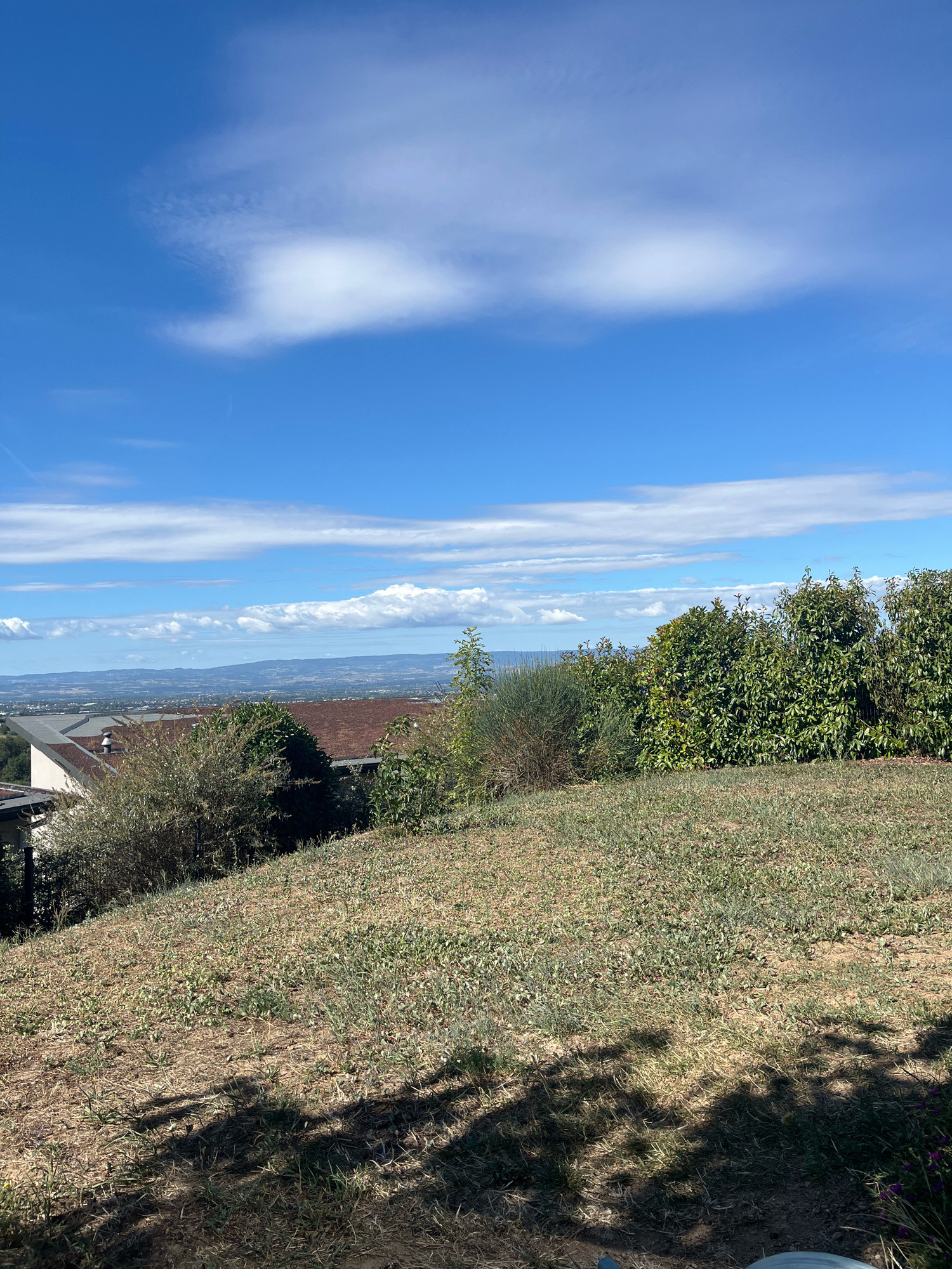 The image depicts a panoramic view from a hillside overlooking the town of Chambœuf, France. The foreground is dominated by a sun-drenched, dry, grassy slope casting long shadows from unseen trees. In the mid-ground, a portion of a building with a red-tiled roof and white siding is visible, partially obscured by lush green bushes and trees. Beyond the immediate vegetation, the town of Chambœuf stretches out, with more buildings and greenery extending towards a hazy, distant landscape. The sky above is a vibrant blue, punctuated by scattered white clouds, with a few wispy formations stretching horizontally across the frame. The overall impression is one of a clear, sunny day in a rural or semi-rural setting.