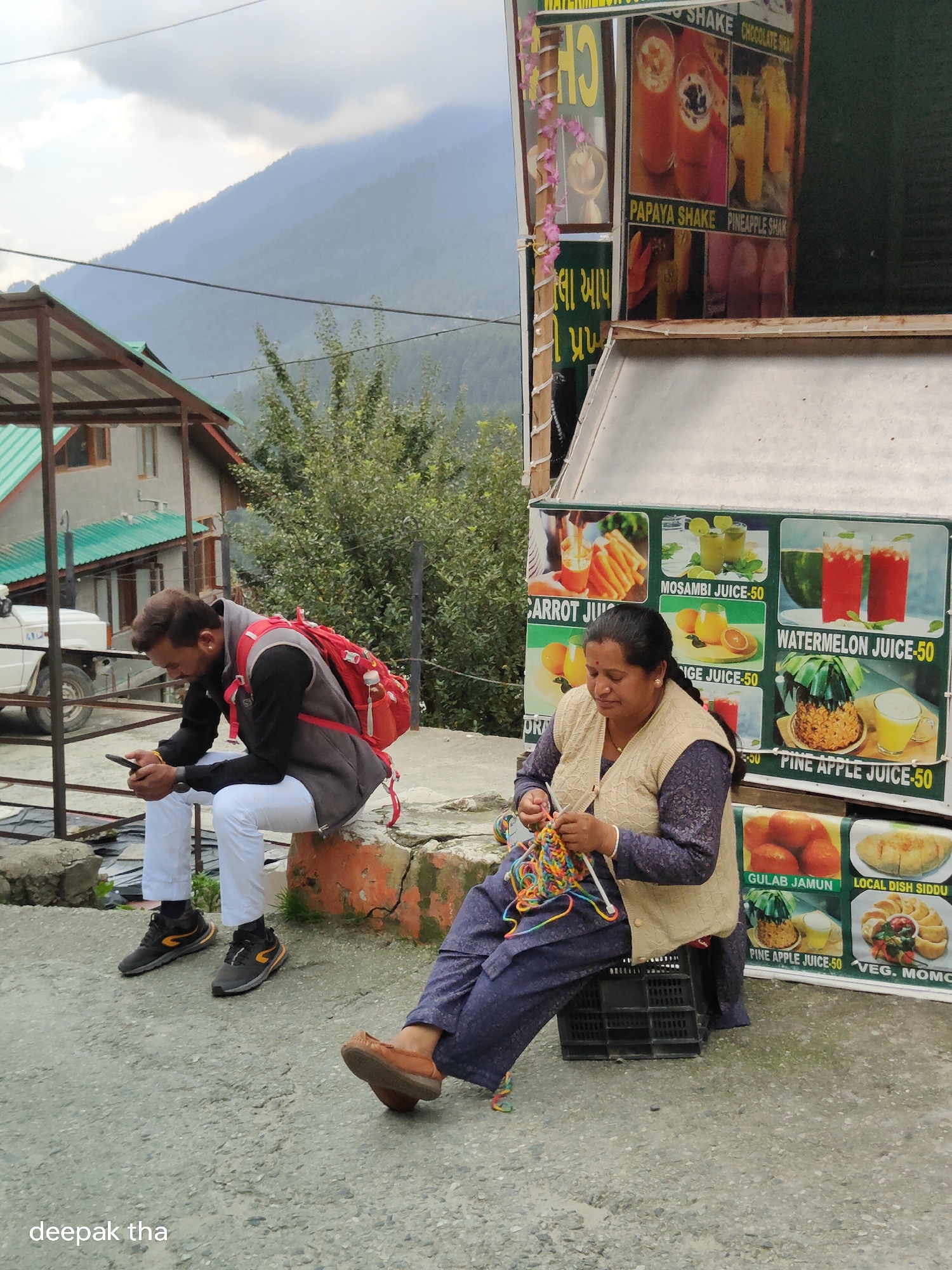 In Vashisht, India, a man wearing a grey vest over a black long-sleeved shirt and white pants sits on a stone ledge, looking at his phone. He has a red backpack with straps over his shoulders, and a water bottle in a side pouch. Beside him, a woman in a beige cardigan over a dark patterned top and blue pants is knitting with multicolored yarn. She is seated on a black plastic crate. Behind them, a food and juice stall displays various drinks and food items, including "PAPAYA SHAKE," "PINEAPPLE SHAKE," "CARROT JUICE," "MOSAMBI JUICE," "WATERMELON JUICE," "ORANGE JUICE," "PINE APPLE JUICE," "GULAB JAMUN," "LOCAL DISH SIDDU," and "VEG. MOMO." The stall is adorned with colorful garlands. In the background, a steep, tree-covered mountain rises under a cloudy sky. A building with a green roof and a white vehicle are visible to the left. The ground is paved with asphalt.