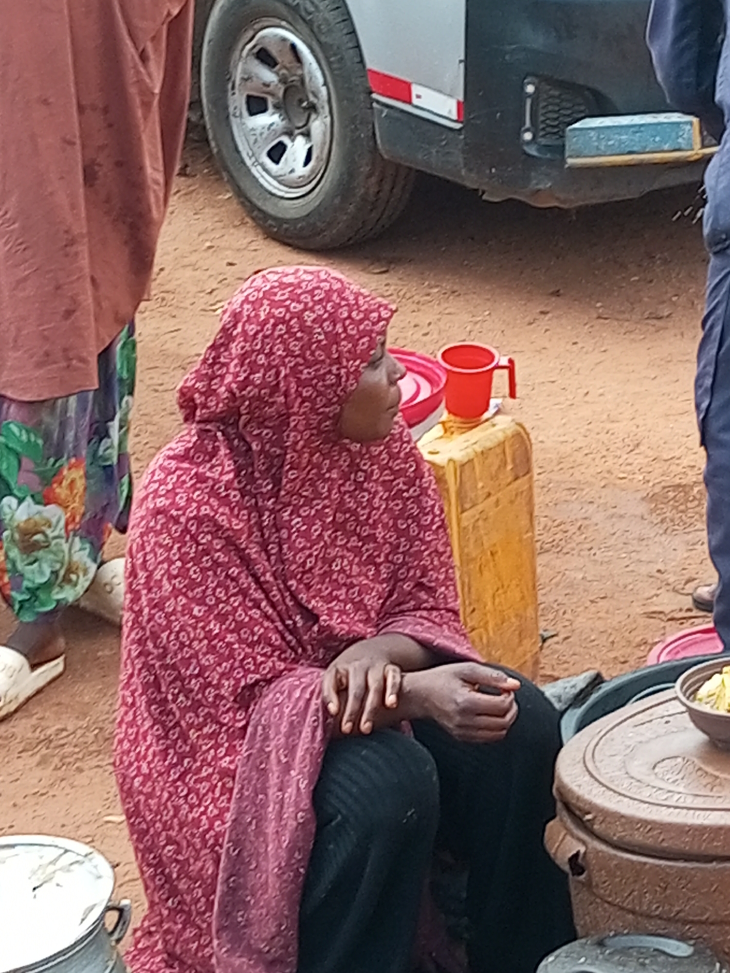 The image depicts a scene on a dirt ground, likely in the city of Gombe, Nigeria. In the foreground, a woman wearing a red hijab with a white floral pattern and a matching patterned garment is seated. Her hands are clasped in her lap, and she is wearing dark pants. To her left, another person, mostly obscured, is wearing a brown garment and a colorful skirt with floral patterns. A white sandal is visible on their left foot.
Behind the seated woman, a yellow jerrycan stands upright, topped with a red plastic bowl and a red mug with a handle. A vehicle is partially visible in the background, including its silver body and a large wheel. The vehicle has a black bumper with some reflective strips. Further to the right, a dark blue-clad person is partially visible, with their lower legs and feet on the ground.
In the lower right of the image, a large, round, brown plastic container with a textured lid is prominent. A smaller metallic pot with a handle is situated in the bottom left corner, and a bowl of food is placed on top of the brown container. The environment appears to be outdoors, with loose soil and scattered debris.