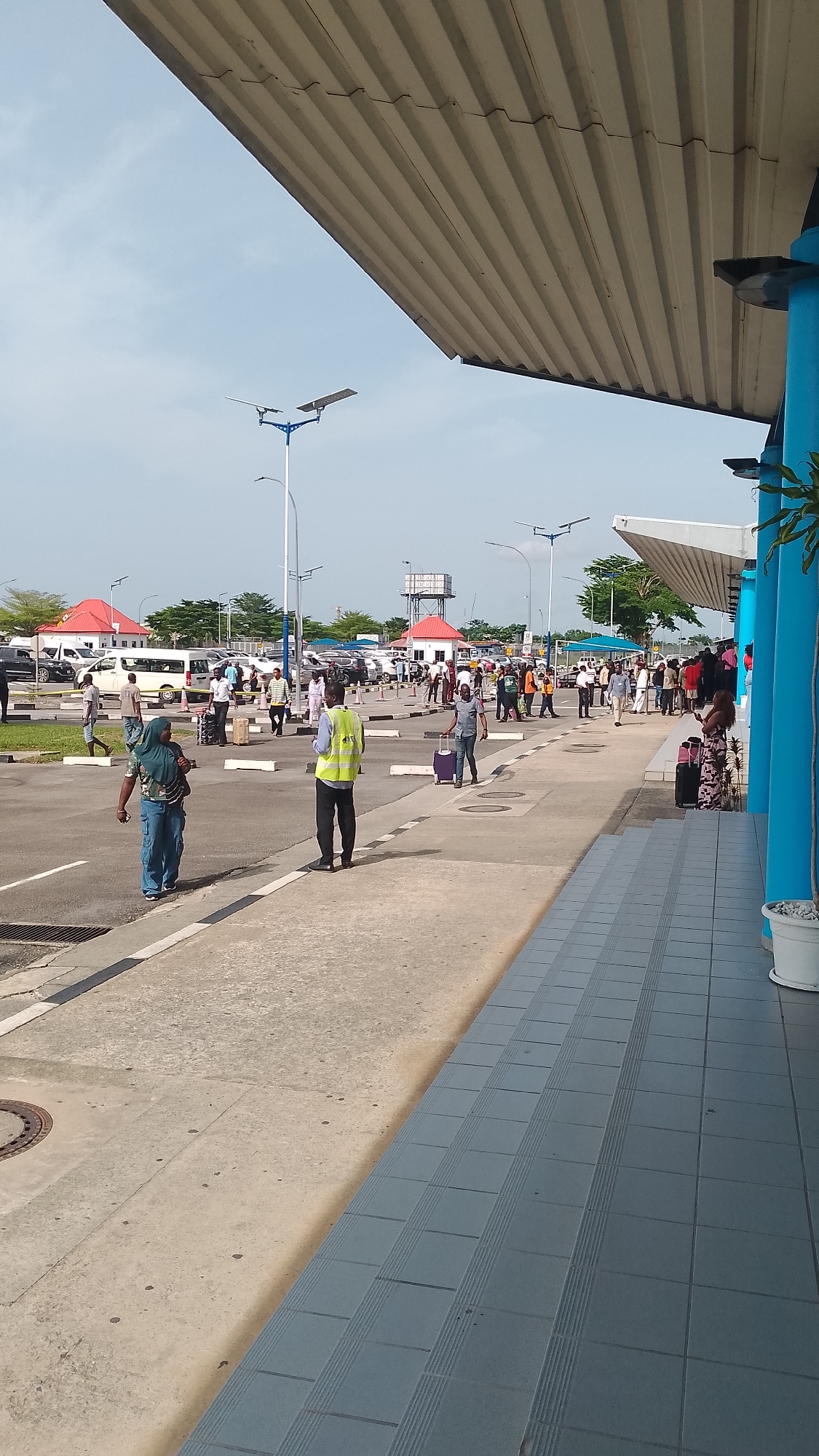 The image shows a scene at an airport or transport hub in Orerokpe, Nigeria. Passengers are walking along a sidewalk area in front of a building with blue columns. Some are pulling luggage. An individual wearing a reflective vest stands in the foreground. In the background, there is a parking lot, vehicles, and small red-roofed structures. Light posts with twin lamps are visible. The sky is partly cloudy.