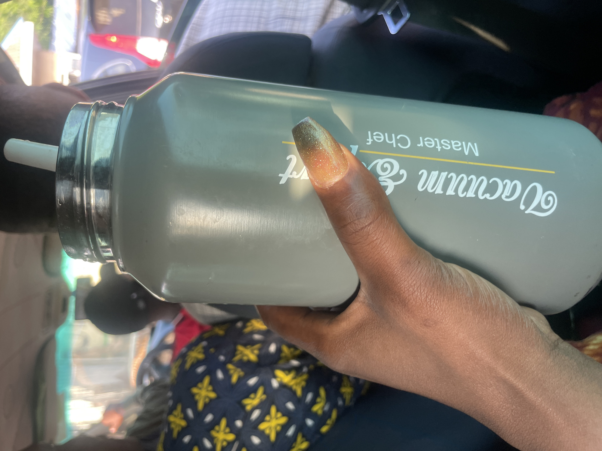 A person's hand with a glittery nail is holding a tall, gray, matte water bottle with a straw inside. The water bottle has "Vacuum & Master Chef" printed on it in white and yellow text. The scene appears to be inside a vehicle, with other people visible in the background, some wearing patterned clothing. The environment suggests an outdoor setting, possibly in Wauro Jabbe, Nigeria, given the lighting and the presence of vehicles.