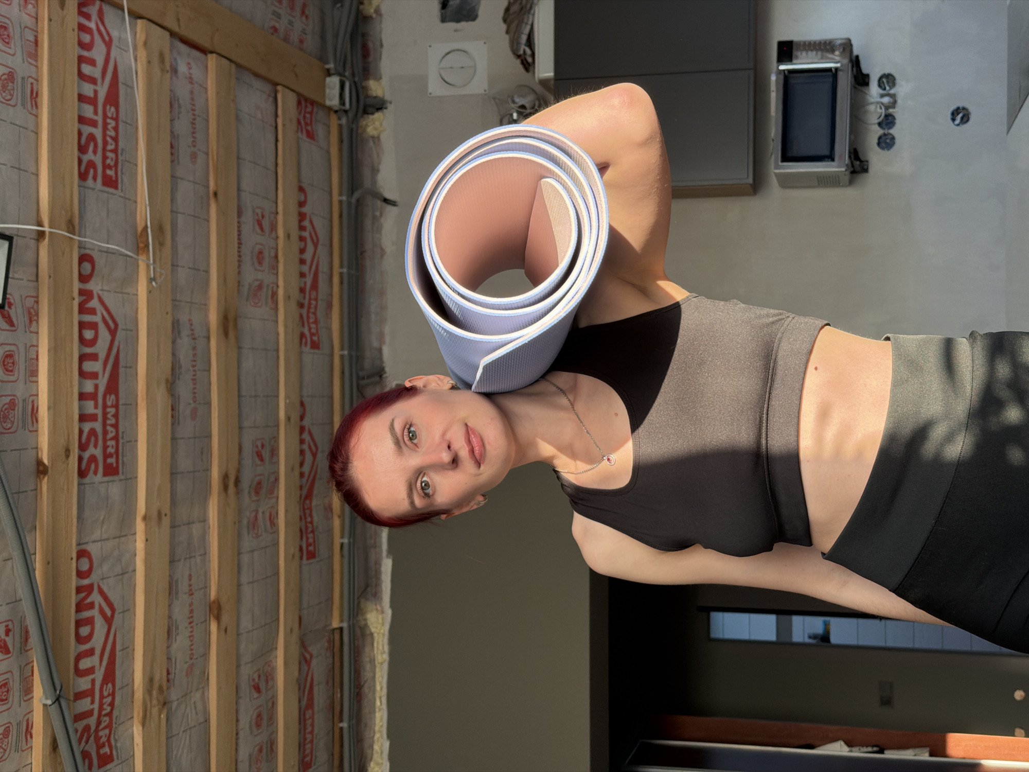 The image, correctly oriented, depicts a woman standing indoors, holding a rolled-up yoga mat. She has red hair, light-colored eyes, and a fair complexion, looking directly at the camera with a neutral to slightly smirking expression. She is wearing a black sports bra or tank top and black athletic bottoms, with a delicate silver necklace visible around her neck. The yoga mat, light blue on its exterior and light pink on its interior, is casually resting on her right shoulder.
The setting appears to be a residential interior in Chaikovsky, Russia, showcasing a blend of ongoing construction and more finished areas. To the woman's left, a wall framing is visible, constructed with vertical wooden studs. Behind these studs, a vapor barrier or insulation material is present, featuring the red text "ONDUTISS," "SMART," and "ondutiss.pro" repeatedly. Exposed electrical wires and conduits are also evident on this unfinished section. To her right, the background shifts to a more completed wall, where a compact metal appliance, possibly an oven or microwave, is mounted. Adjacent to it are several electrical outlets and wiring. Below this, a darker, presumably finished area of the room or a connecting hallway is visible, with a window offering a blurred view of the outdoors.
The lighting is bright, casting strong, defined shadows on the woman and the walls, indicative of direct sunlight, suggesting it is midday or early afternoon on a clear day. The overall scene captures a moment likely before or after a fitness activity, set against the backdrop of a home that is either being renovated or is in various stages of completion.