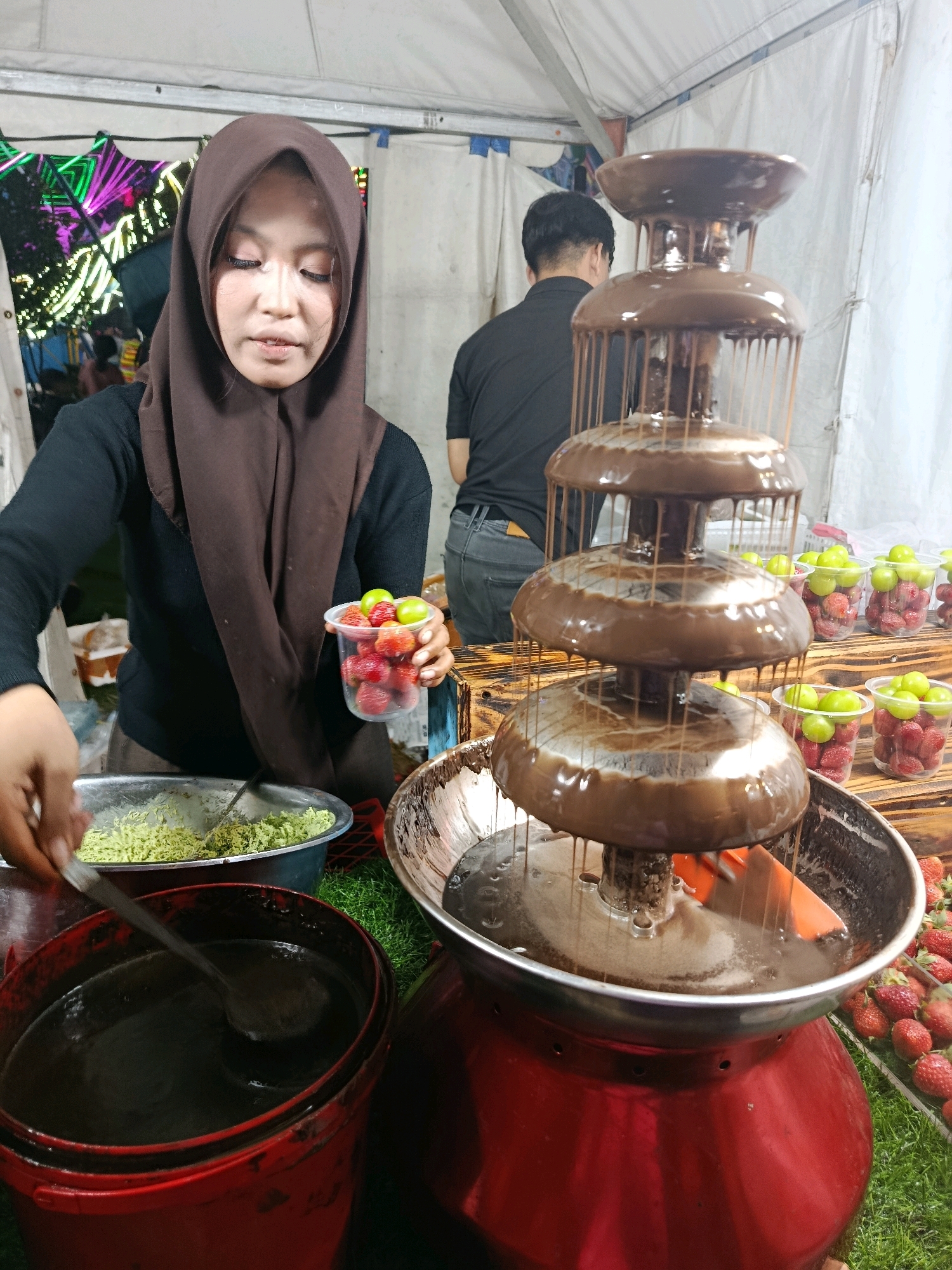 In Balaraja, Indonesia, a woman wearing a brown hijab and a black long-sleeved shirt is preparing a chocolate fountain. She holds a clear plastic cup filled with strawberries and green grapes in her right hand. Her left hand is reaching into a large metal bowl containing a light green, shredded substance, possibly pandan, holding a spoon. A red bucket with a dark liquid and a ladle sits to her left. In front of her is a three-tiered chocolate fountain, with melted chocolate cascading down each level. The fountain is placed on a red base. To the right of the fountain are clear plastic cups filled with strawberries and green grapes, arranged on a wooden surface. In the background, another person in a black shirt is visible. The scene appears to be set up outdoors, under a white tent, with colorful lights in the far left background.