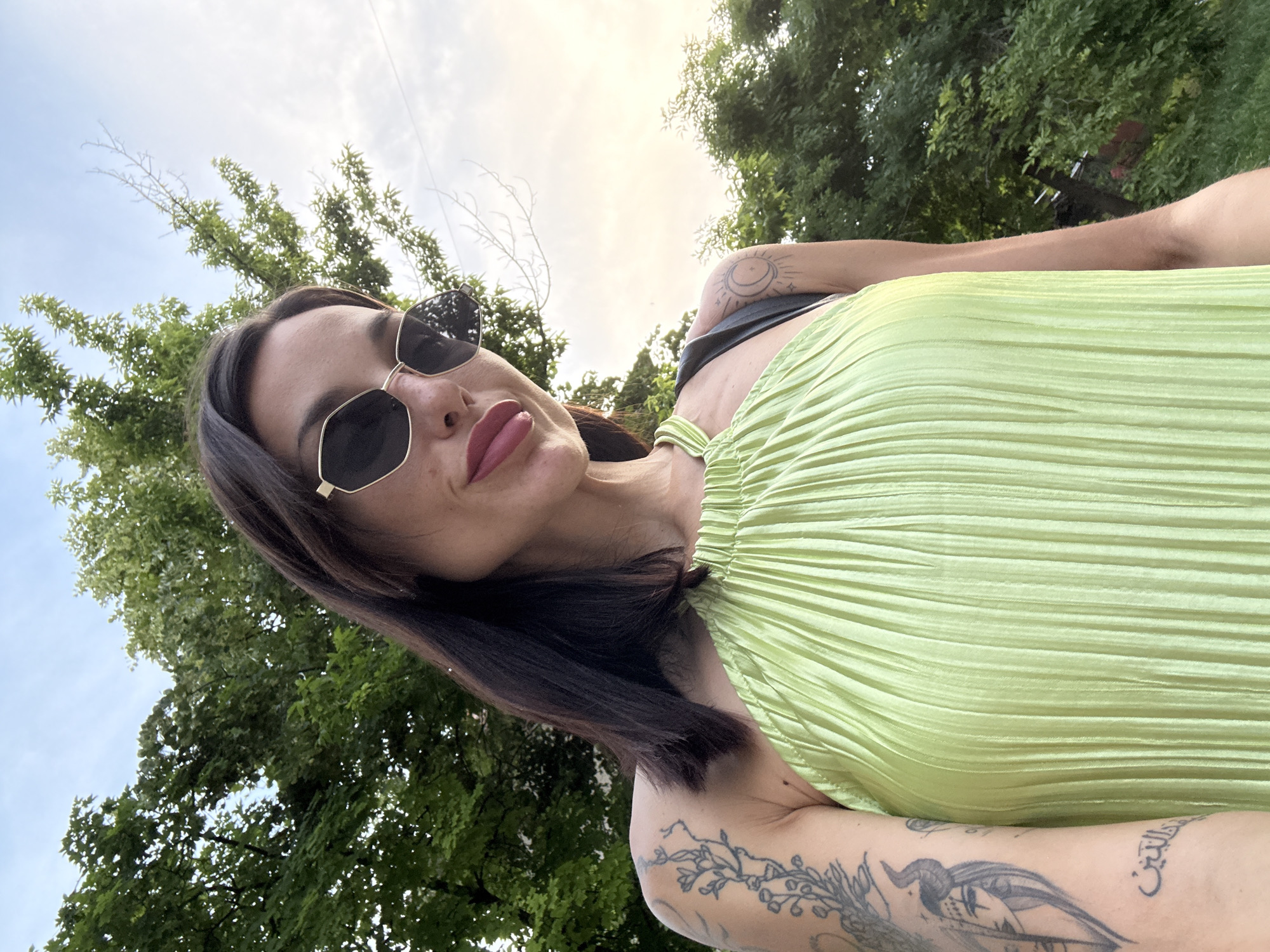 A woman is taking a selfie outdoors. She has dark hair, sunglasses, and is wearing a green dress. She has tattoos on her arm. She is standing in front of trees and a cloudy sky. The location is not specified in the image or the prompt.