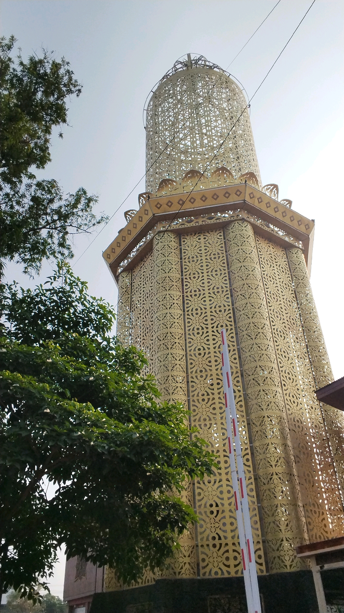 The image captures a low-angle view of a tall, ornate golden tower with intricate geometric cutouts, possibly a minaret or a decorative structure. The tower is situated in Mojokerto, Indonesia, as indicated by its architectural style. In the foreground, a lush green tree with dense foliage partially obscures the lower portion of the tower. A white and red striped barrier arm is visible, suggesting it might be an entrance to a facility. The sky is a pale, overcast blue, with a slight haze or lens flare adding to the brightness of the upper portion of the frame. The overall impression is of a sunlit day, with the golden tower prominently displayed against the sky, framed by the natural element of the tree.