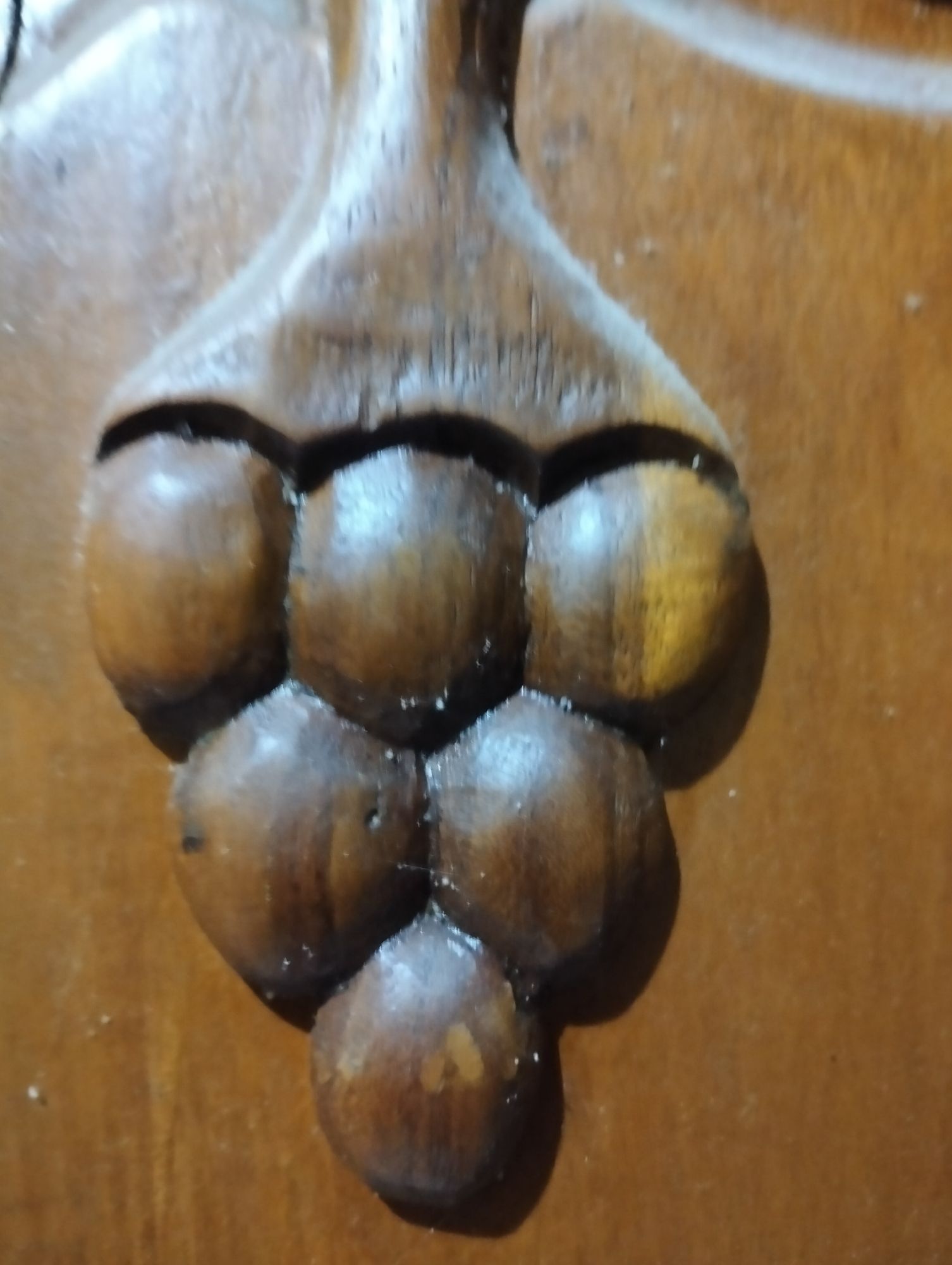 A close-up, low-angle shot shows a carved wooden decoration that resembles a bunch of grapes. The carving is detailed, with individual grape-like forms clustered together and attached to a central stem-like structure. The wood has a warm, brown hue and appears to be slightly glossy, suggesting a finish. The background is a flat, textured wooden surface. The image does not contain any information that allows for the determination of its location in the city of Payagaboh, Indonesia.