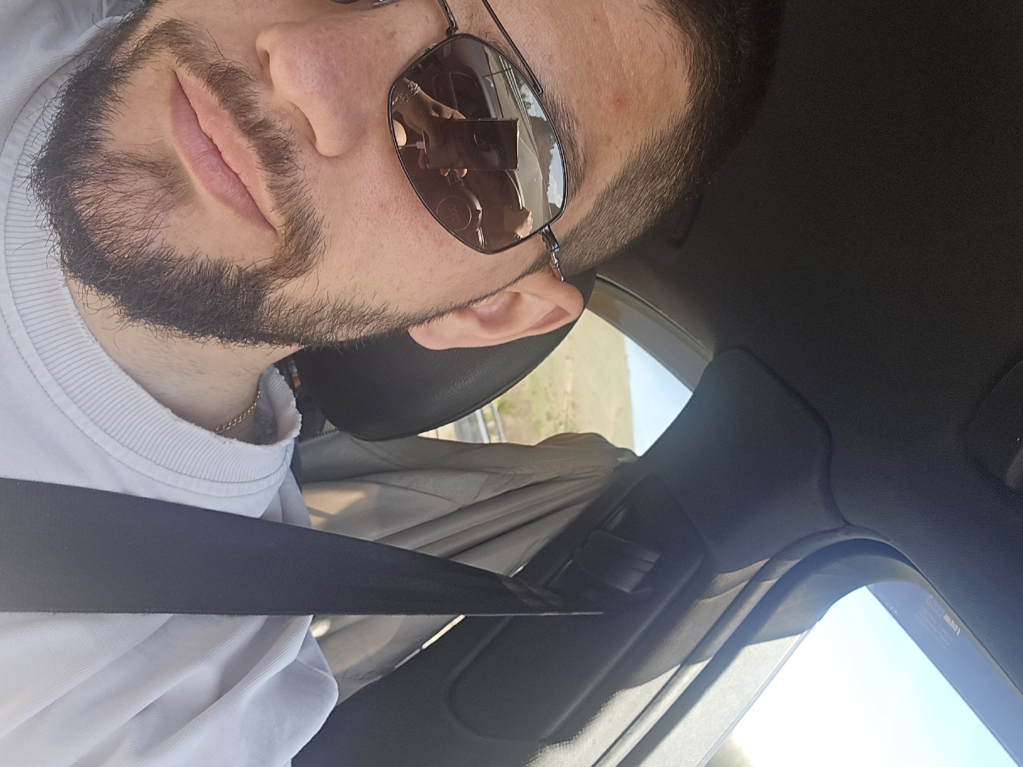 The image is a selfie taken from inside a car. A man with a beard and sunglasses is visible, wearing a white shirt and a seatbelt. The reflection in the sunglasses shows a phone and headphones. The background shows an outside view of land and sky. There is no indication this photo was taken in Staro Selo, Bulgaria.