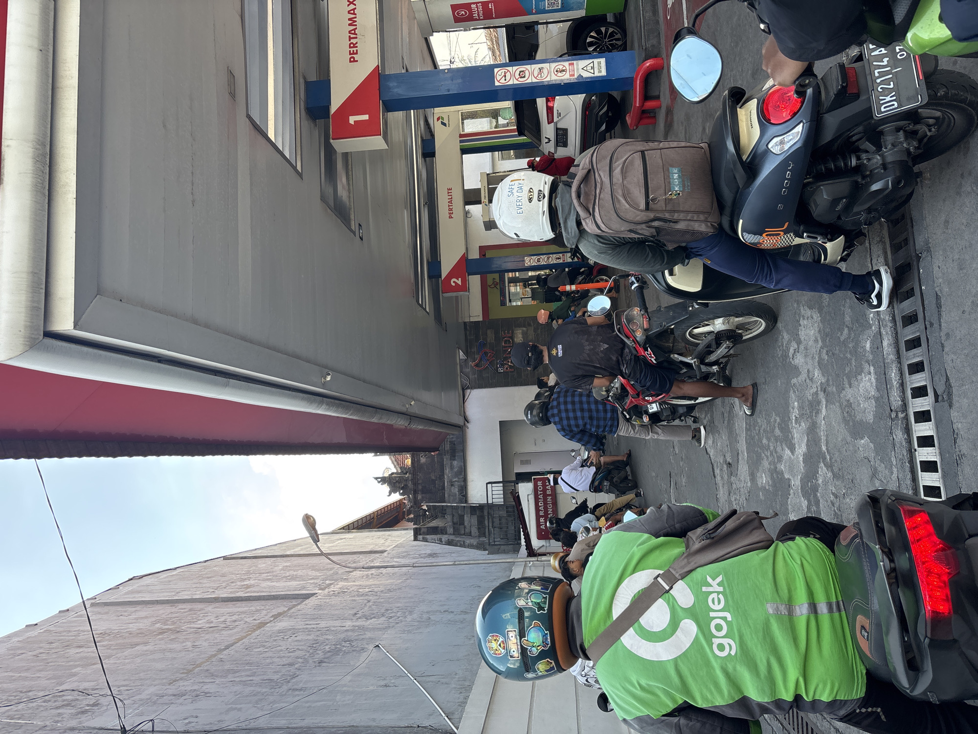 A line of motorcycles and a white SUV are waiting at a Pertamina gas station in Denpasar, Indonesia. A man in a bright green Gojek jacket is in the foreground, seated on a motorcycle. Another person wearing a white helmet with "BE SAFE EVERY DAY" written on it is behind him, also on a motorcycle. The gas station has pumps labeled "PERTAMAX" and "PERTALITE." The sky is bright and clear.