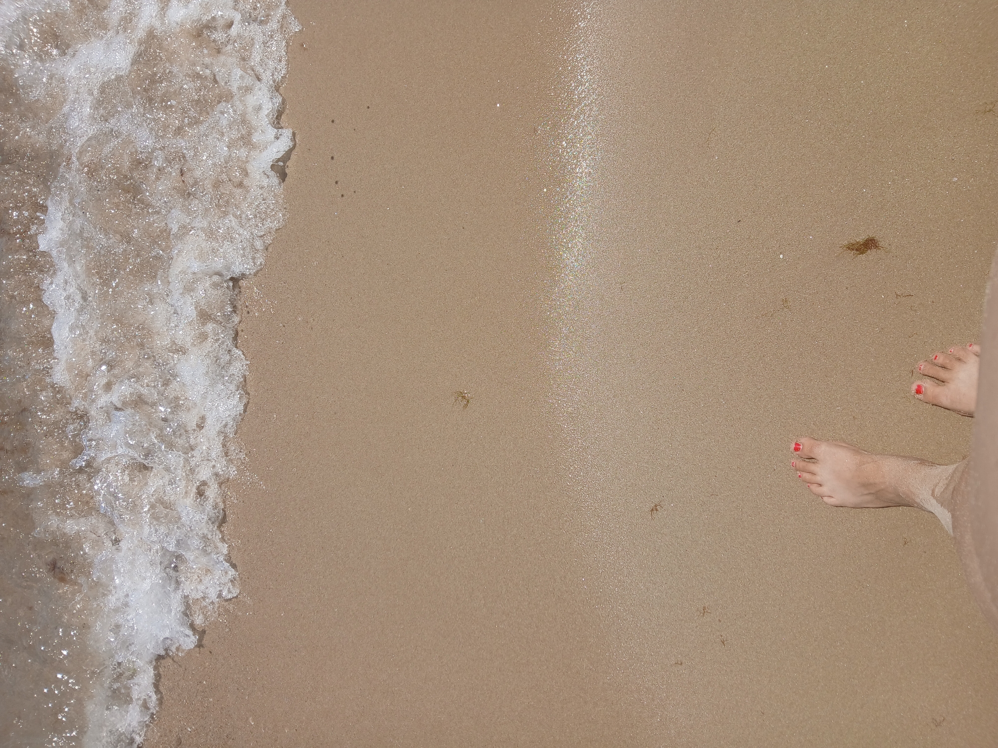The image is an overhead shot of a sandy beach in Kranevo, Bulgaria, with the edge of the sea encroaching on the shore. Two bare feet, with red nail polish on the toes, are visible in the lower portion of the frame, suggesting the viewer is standing on the beach. The sand is light brown and appears wet near the water's edge, which is a foamy white wave. There are small pieces of seaweed and sand debris scattered across the beach. The overall scene depicts a tranquil beach environment.