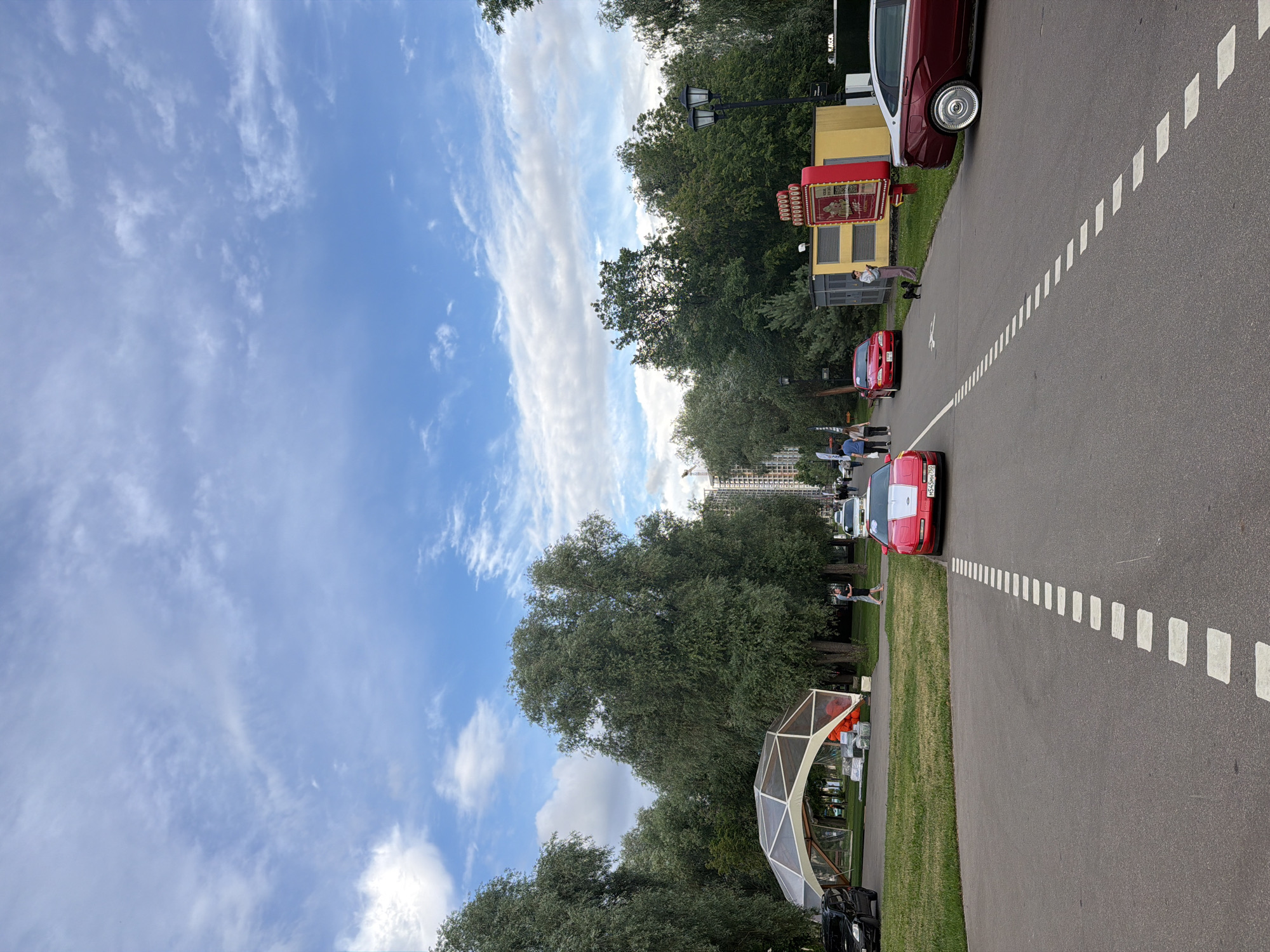 The image depicts an outdoor scene with a paved road in the foreground, marked with dashed white lines. Several cars are present on and near the road, including a red sports car, a red sedan, and a black SUV. To the left of the road, there's a grassy area with a large, geodesic dome-like structure. Beyond the dome, a line of trees forms a backdrop, and in the distance, several tall buildings are visible, suggesting an urban or suburban environment. The sky is blue with scattered white clouds. A few people are scattered throughout the scene, some walking near the cars and others near the dome. There's also a lamppost and a yellow structure with a red sign.