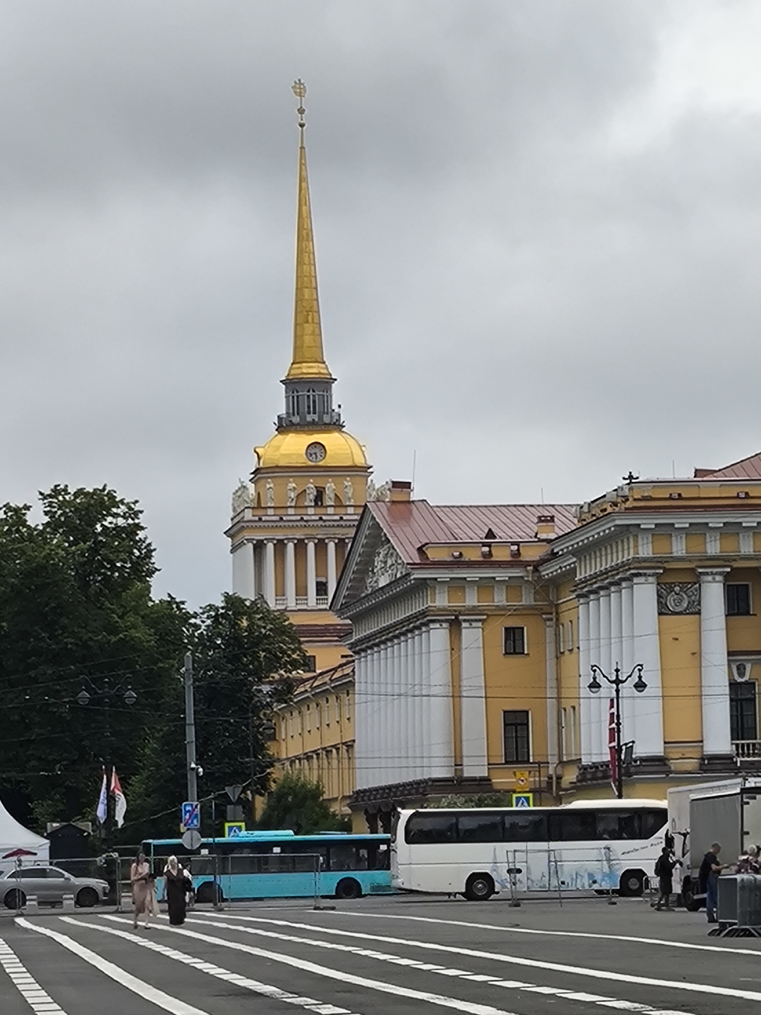 The image captures a view of the Admiralty Building in Saint Petersburg, Russia. Dominating the skyline is the building's iconic golden spire topped with a ship weather vane, set against a cloudy sky. In the foreground, a wide street with a pedestrian crossing is visible, with several vehicles present. A bright turquoise bus is positioned on the left, and a white tour bus is further to the right. Several people are seen on the sidewalk and crossing the street, some appearing to be tourists. The yellow facade of the Admiralty Building, with its classical columns and statues, occupies the mid-ground, partially obscured by lush green trees. The overall scene depicts a typical urban streetscape in a historic European city on an overcast day.