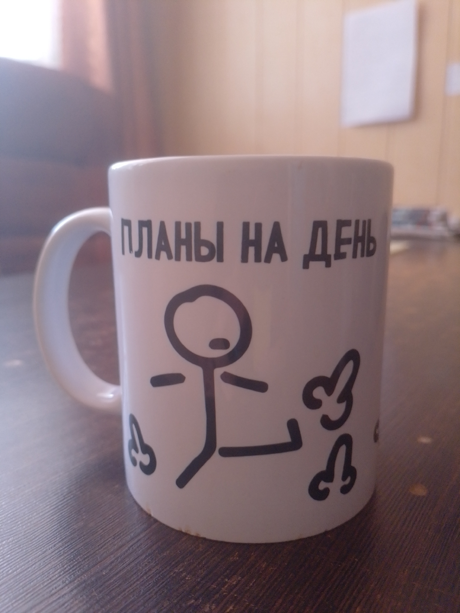 A white ceramic mug with black lettering and a drawing sits on a dark wooden surface. The lettering on the mug reads "ПЛАНЫ НА ДЕНЬ" (Plans for the day). The drawing is a stick figure with a round head, a horizontal line for arms, and a vertical line for a body. There are also drawings of what appear to be phalluses on the mug. The background is out of focus, showing a warm-toned wall and a glimpse of what might be furniture. There are no people visible in the image. The overall lighting suggests an indoor setting with natural light coming from the left.