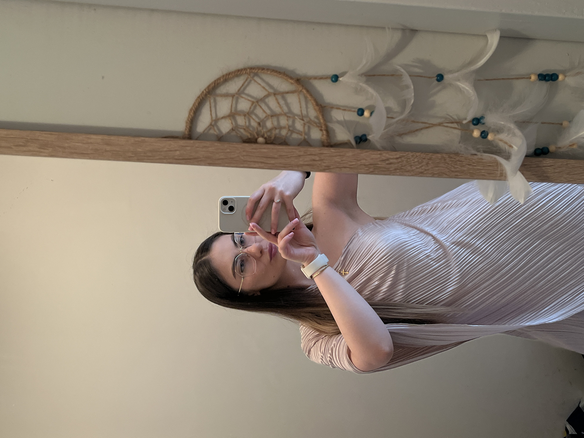 A woman is taking a selfie in a mirror. She is wearing glasses, a white smart watch, a ring, and a light pink pleated dress. She is holding up a white smartphone with her left hand, making a peace sign with her right hand. Above the mirror is a decorative dreamcatcher with feathers and beads. The wall behind the mirror is a pale beige color. The location is indoors and appears to be a private residence in the city of Unknown, Unknown.