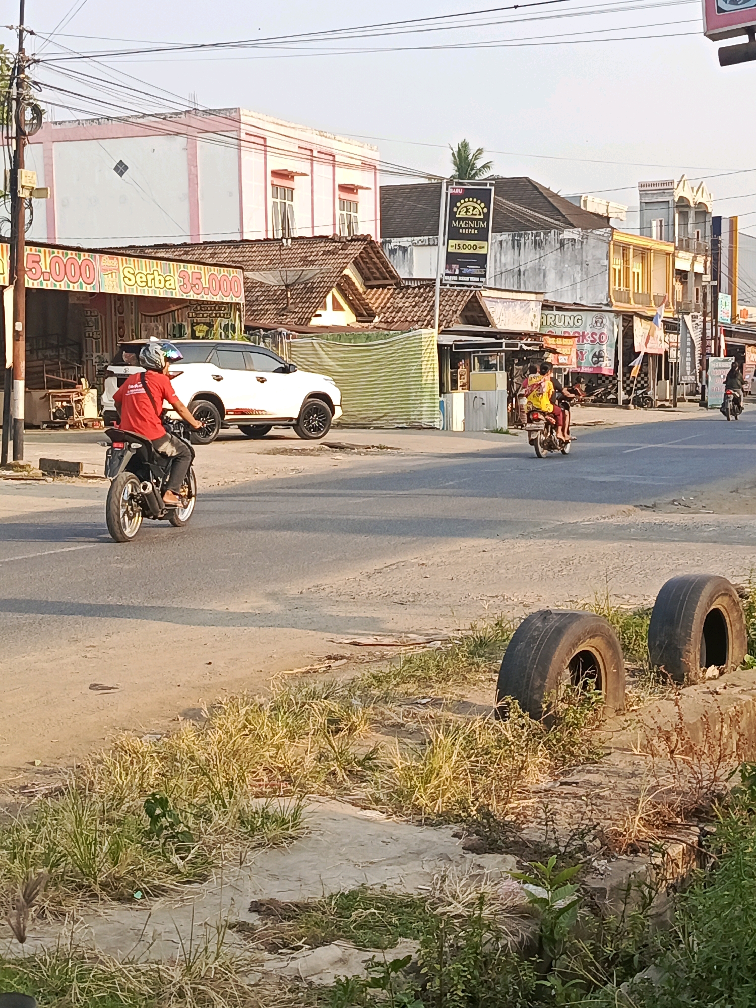 The image depicts a street scene in Megang Sakti II, Indonesia. The foreground shows dry grass and two discarded tires. In the midground, a person wearing a red shirt and black pants rides a motorcycle in the right lane of a paved road. The person is wearing a helmet. A white SUV is parked on the left side of the road. In the background, buildings line the street, featuring signs with text in Indonesian. The sky is clear and bright, indicating daytime. Electrical wires are visible overhead.