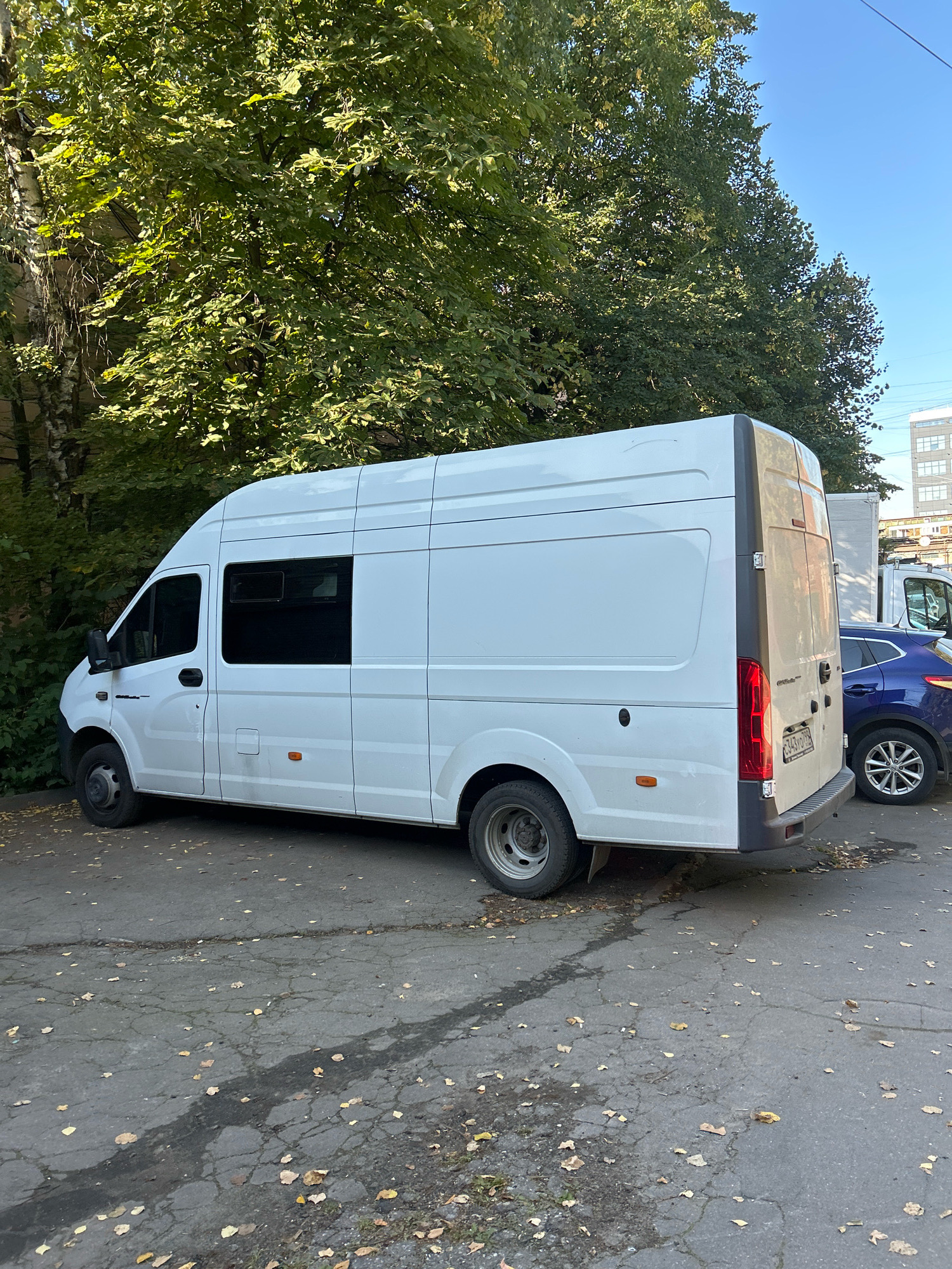 A white commercial van is parked on a cracked asphalt surface in an urban setting. The van, a Gazelle Next model, is oriented to the left, with its rear facing the viewer. It has tinted windows along its side and a dark grey vertical trim piece along the rear door. The license plate is visible on the rear of the van and reads "C343YO 199". To the right of the van, a dark blue hatchback car is partially visible. A large tree with green foliage overhangs the scene from the left, casting shadows on the ground. The ground is littered with fallen yellow and brown leaves. In the background, there are buildings and power lines, suggesting a city environment, possibly Moscow, Russia, given the Cyrillic on the license plate. The sky is clear and blue.