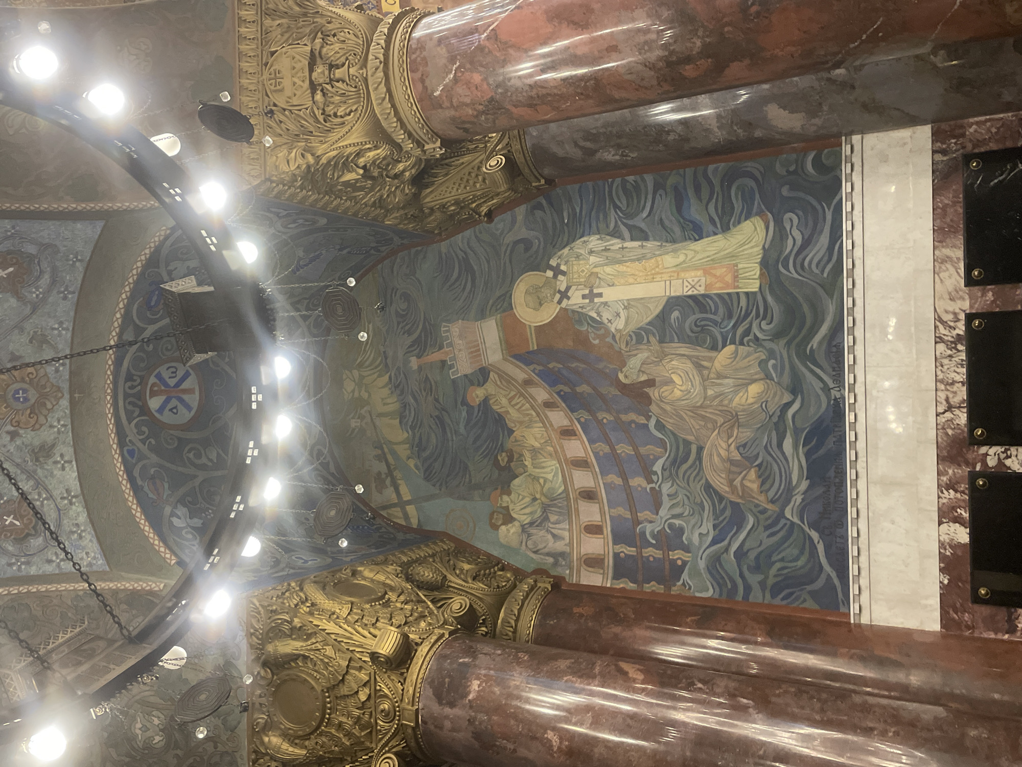 The image shows an interior view of a church, likely within the city of Kronstadt, Russia, featuring a large mural and ornate architectural elements. The mural dominates the central portion of the frame, depicting a scene with figures in a boat amidst stylized waves. One prominent figure, appearing to be a saint in religious vestments with a halo, is reaching down towards another figure kneeling in the water. A boat, likely representing Noah's Ark, is visible behind them with multiple figures on its deck. The mural is rendered in a traditional, possibly Byzantine, style with rich colors and flowing lines.
Flanking the mural are two large columns, one on the left and one on the right. These columns are made of dark, possibly red, marble with a polished finish, and are topped with elaborate golden capitals adorned with intricate carvings. Above the mural, the ceiling is also decorated with colorful frescoes and architectural details, including chains and lighting fixtures. The overall impression is one of grandeur and religious solemnity, characteristic of historic Russian Orthodox churches. The lighting in the image is bright, with several spotlights illuminating the scene.