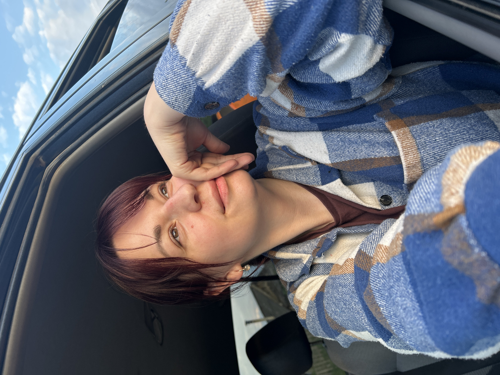 A young woman with reddish-brown hair is sitting in the driver's seat of a vehicle, looking out the open window to her right. Her right hand is propped against her chin, and her left arm rests on the car door. She is wearing a blue, white, and brown plaid shirt over a maroon t-shirt. The background shows a cloudy sky and hints of rural landscape, suggesting a location outside of a dense urban area. The photo appears to have been taken outdoors during the daytime.