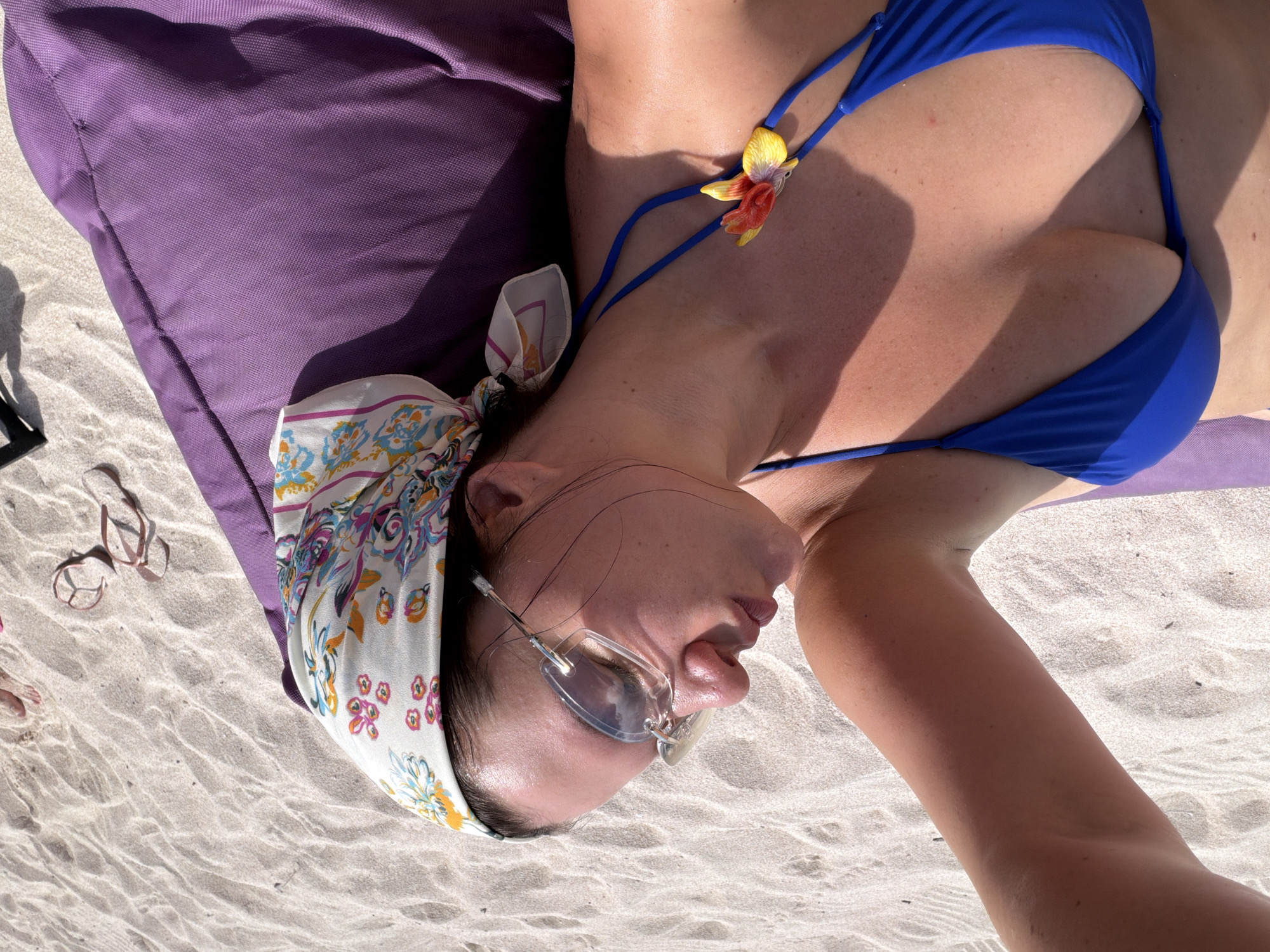 A woman lies on a sandy beach, resting on a purple mat, wearing a blue bikini and a patterned headscarf. She has on clear-rimmed sunglasses and her eyes are closed, her head tilted back. Sunlight illuminates her skin, creating shadows across her face and body. A decorative yellow and red flower adorns the string of her bikini top. In the background, the sand is rippled, and a pair of light pink flip-flops are visible. The image captures a moment of relaxation on what appears to be a sunny day on a beach in Nusa Penida, Indonesia.