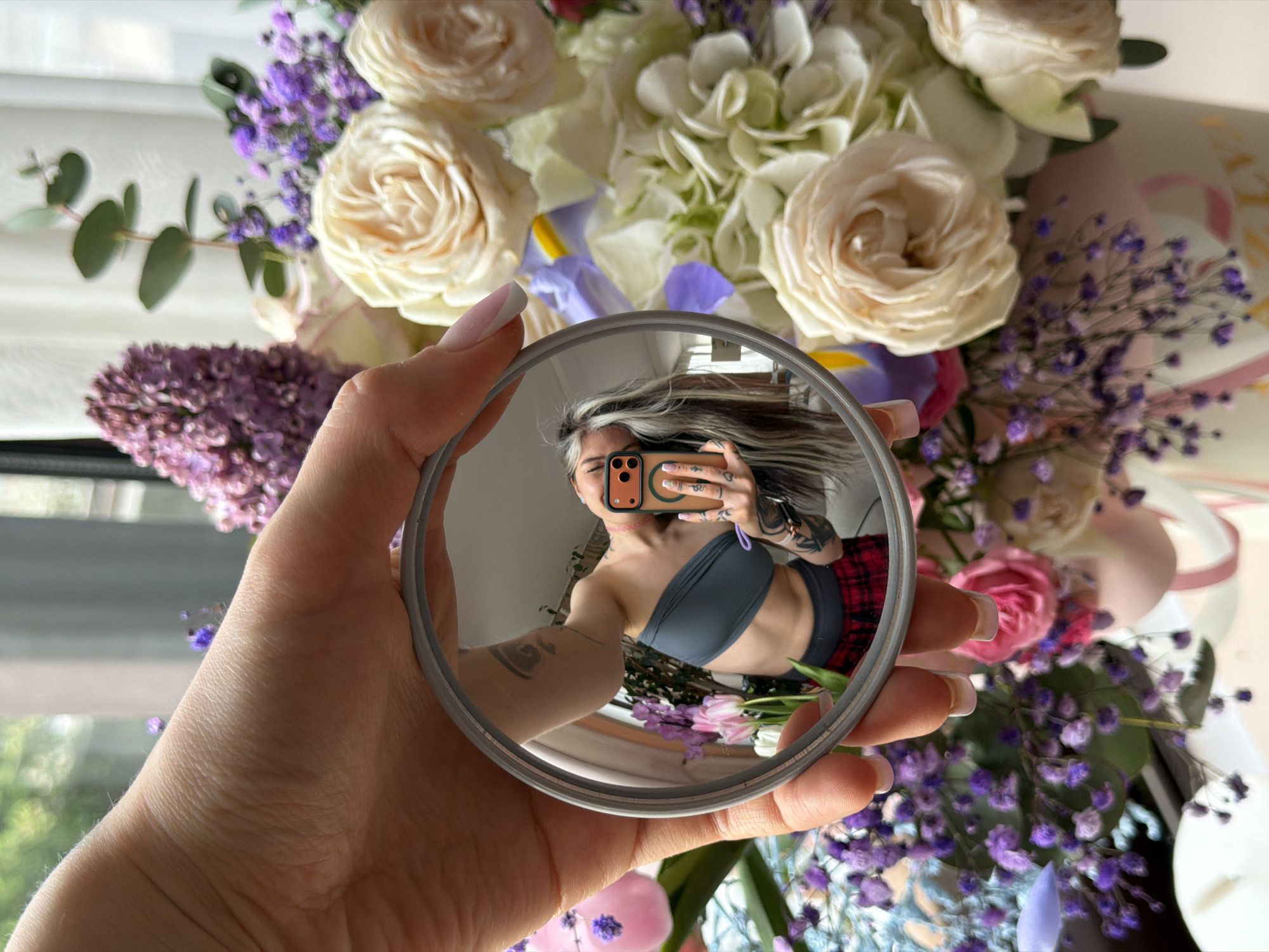 A hand holds a round mirror in front of a bouquet of flowers. The reflection in the mirror captures a person taking a selfie with a smartphone. The person is wearing a grey strapless top and red and black plaid pants. The background through the window appears to be a residential or natural area, with indistinct greenery. The scene is indoors, with ample natural light suggesting it may be near a window. There is no specific evidence to ascertain that the location is Budva, Montenegro.