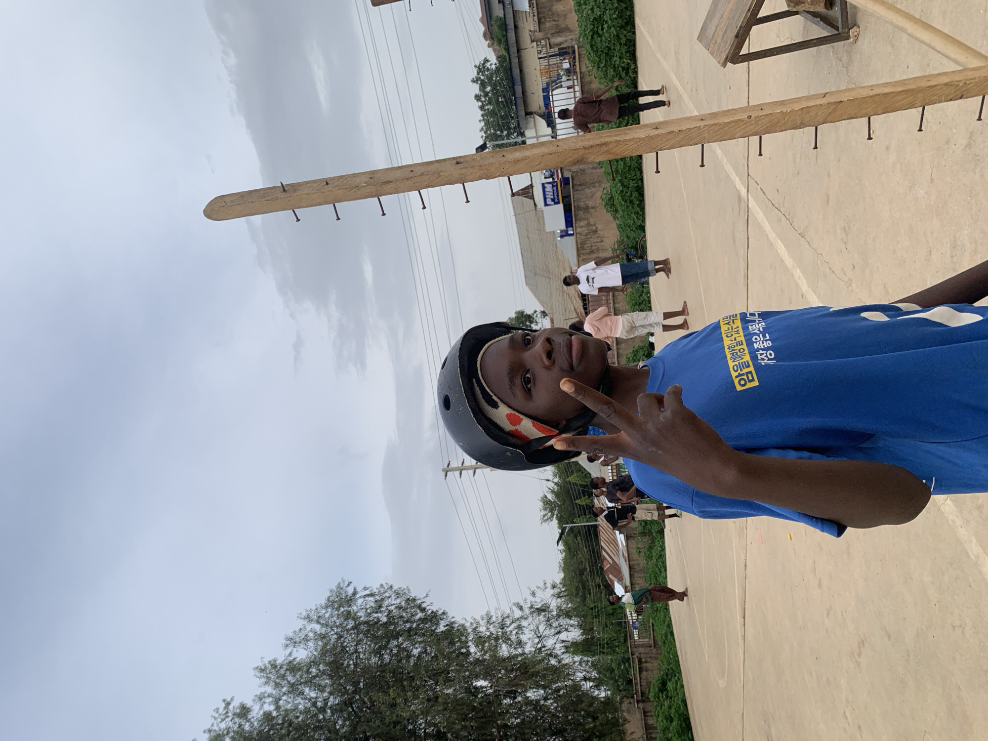 The image shows an individual in a blue t-shirt and black helmet, making a hand gesture. In the background, a wooden horizontal bar with nails is visible, along with several other people. The location appears to be in Jos, Nigeria, based on the presence of distinctive local architecture and vegetation. The ground is concrete, suggesting an outdoor court or open space. Power lines are also present in the background.