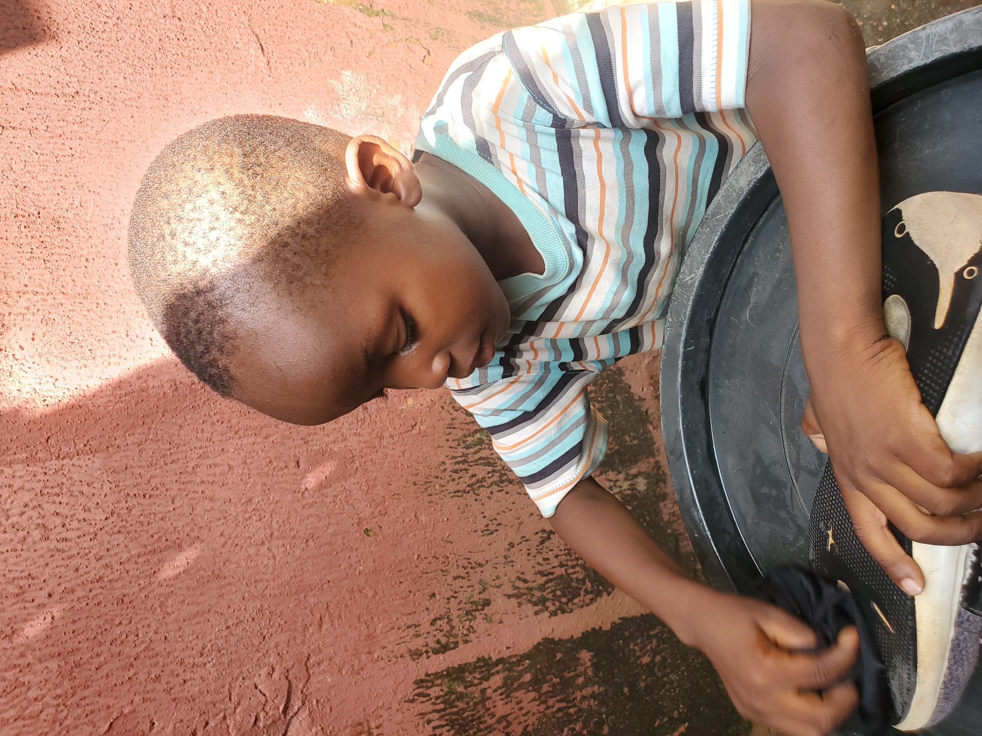 In this overhead shot, a young boy is focused on cleaning a shoe. He is wearing a striped t-shirt with horizontal bands of blue, white, orange, and black. His dark hair is closely cropped, and his eyes are downcast as he concentrates on his task. He is using a black cloth to wipe a black shoe, which appears to be a sneaker with white soles. The shoe is placed in a large, dark-colored basin.
The background consists of a textured, reddish-brown surface that looks like an outdoor wall or ground. There are areas of moss or dampness on this surface, suggesting it might be an outdoor setting. The lighting indicates it is daytime, with sunlight casting shadows. The scene appears to be in Jos, Nigeria. The boy's posture and focused expression convey a sense of diligence and responsibility.