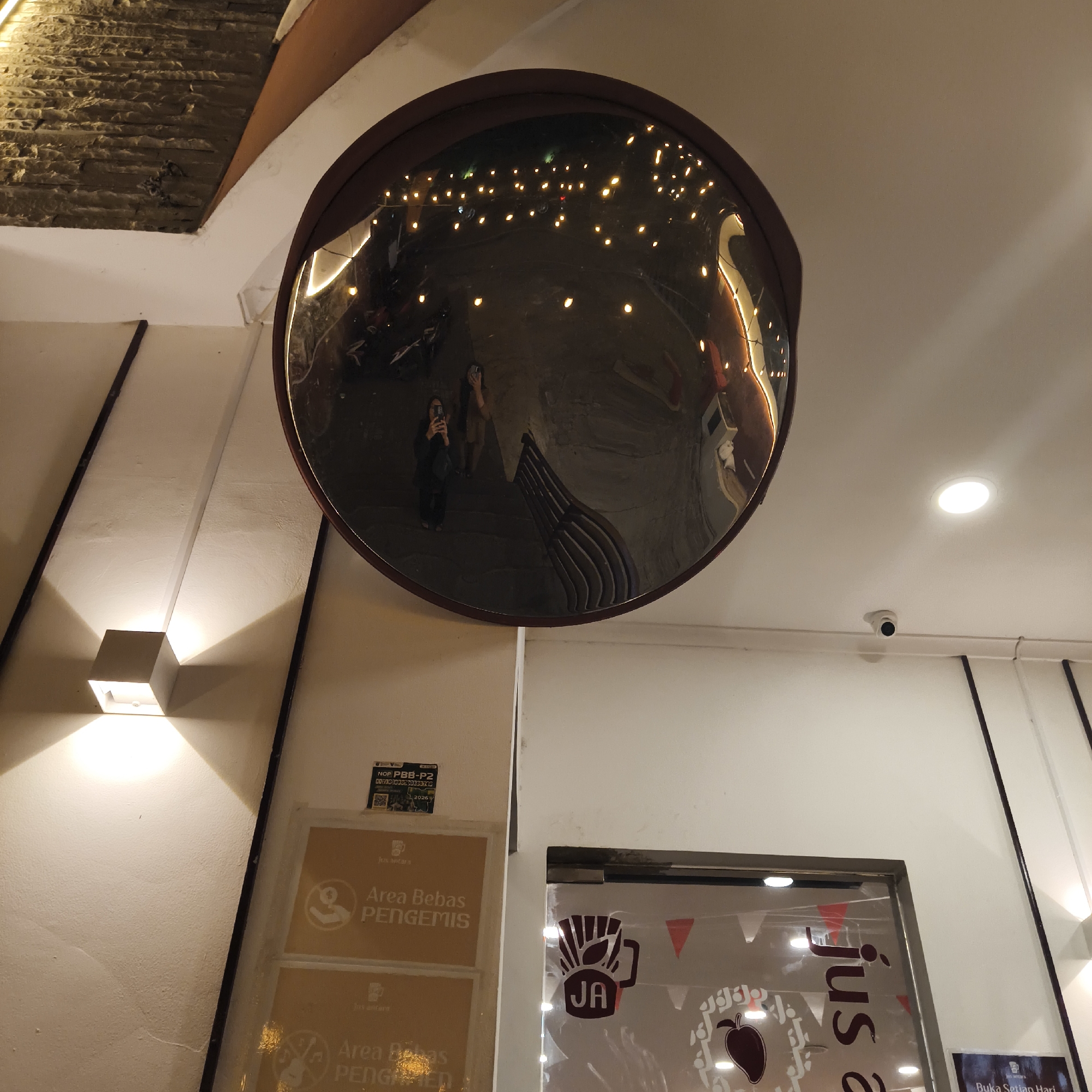 The image shows a wide-angle convex mirror reflecting a street scene. In the reflection, two individuals are visible, one holding a phone as if taking a picture. The mirror is mounted on a white wall, illuminated by a square wall-mounted light fixture below it. To the right of the mirror, a surveillance camera is mounted on the ceiling. Further to the right, a glass-paneled doorway is visible, with decorations and signage, including the text "Jus &". Below the mirror, on the wall, are two yellow signs with text that appears to be in Indonesian, one reading "Area Bebas PENGEMIS" which translates to "Beggar-Free Area". A small sticker with a QR code and some text is also present on the wall. The ceiling is white, with some visible lighting fixtures. The overall setting suggests an establishment, possibly a shop or restaurant, in an urban environment. The presence of the "Area Bebas PENGEMIS" signs suggests a location in Indonesia, where such notices are common in public and private spaces. While the specific location in South Jakarta, Indonesia, cannot be definitively identified from the image alone, the context provided by the signage points to this general region.