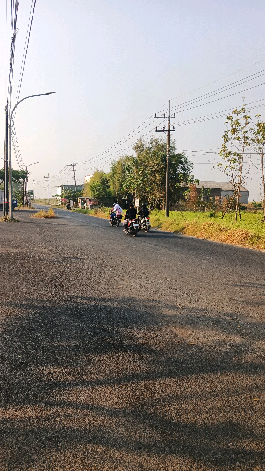 The image depicts a rural road in Ngepung, Indonesia, during daylight hours. A paved road runs diagonally across the frame, with its surface showing shadows cast by overhead trees. Three individuals on motorcycles are traveling away from the viewer down the road. The motorcycle closest to the viewer has a passenger wearing a black shirt and helmet, and the rider is wearing a dark outfit. The middle motorcycle has a rider in a dark outfit and a passenger wearing a white garment. The motorcycle furthest away appears to have at least one person on it, though details are obscured.
To the right of the road, a grassy verge is visible, bordered by trees and a simple building with a weathered, grey roof. In the distance, a cluster of tall bamboo-like plants and other foliage line the landscape. Electrical poles with power lines are scattered throughout the scene, indicating utility infrastructure. The sky above is clear and light blue, suggesting fair weather. The overall impression is that of a quiet, everyday scene on a road outside of a populated area in Ngepung.