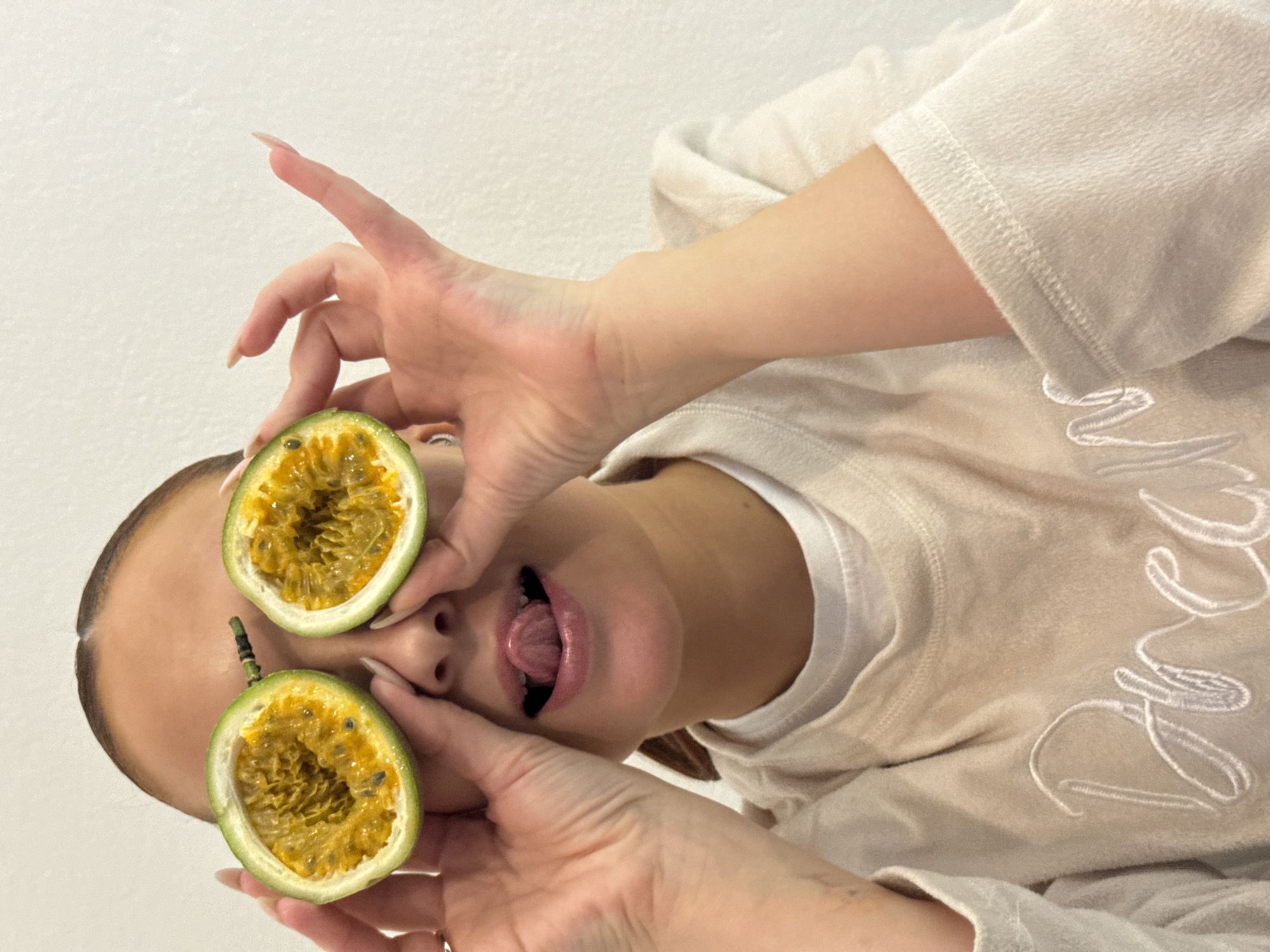 An individual is depicted indoors in Chaikovsky, Russia, against a plain, light-colored wall. The subject, appearing to be a young female, is positioned centrally in a medium close-up. She is performing a playful action, holding two halves of a green passion fruit over her eyes, with the vibrant yellow-orange pulp and dark seeds visible. Her mouth is open, and her tongue is extended slightly. Both hands are visible, positioned at eye level, holding the fruit; the left hand shows a ring on the ring finger, and both hands feature long, manicured fingernails. The subject is wearing a light-colored (beige or cream) long-sleeved top, possibly a sweatshirt, over a white undershirt. Embroidered text, partially visible on the front of the top, spells out "Dream". The lighting is even, illuminating the subject and the immediate background.