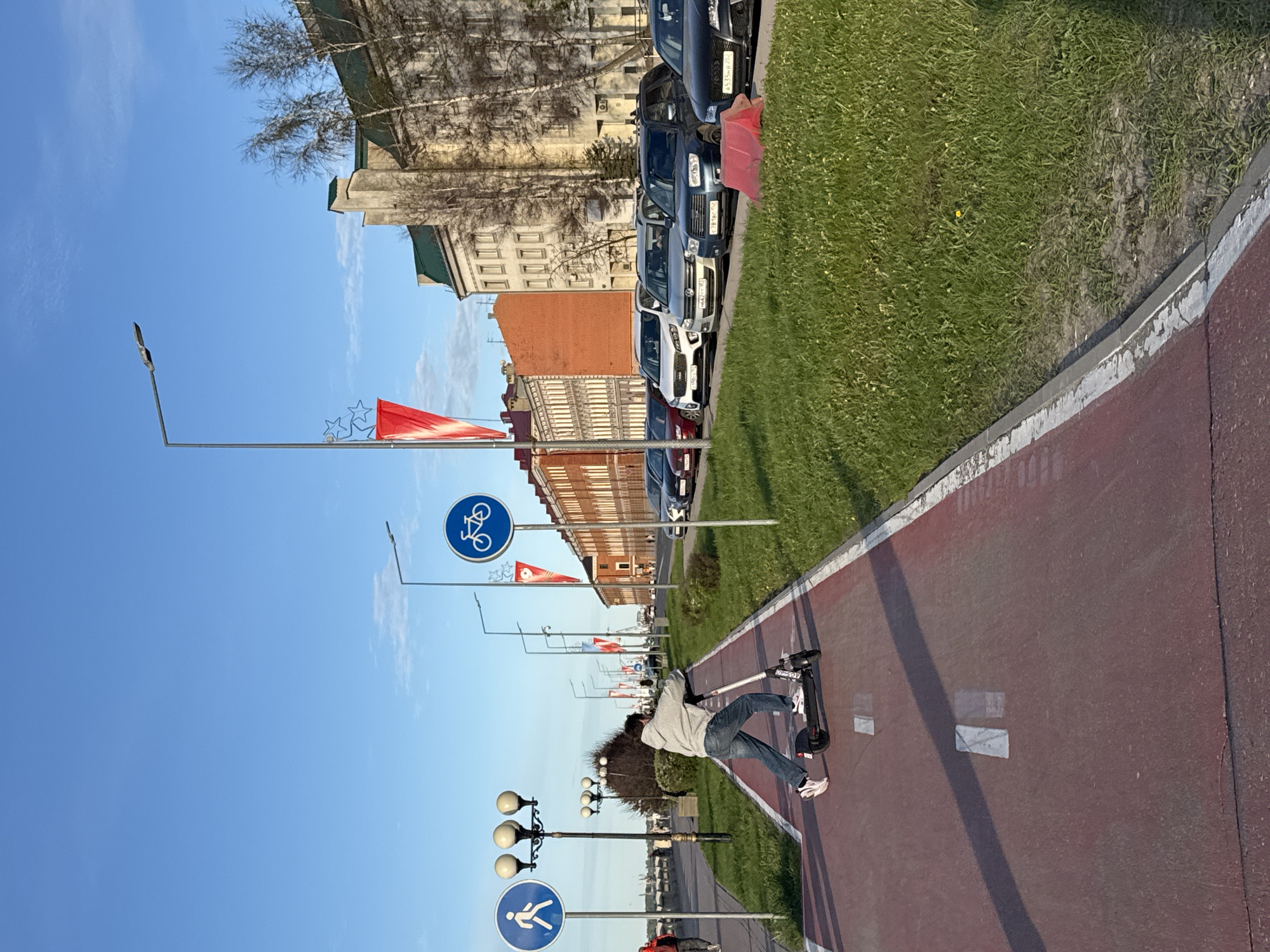 A street scene in Nizhny Novgorod, Russia. A person rides a scooter on a red pathway. A row of parked cars is visible on the right, along with grass and a building. On the left, there are lampposts, signs for bicycle and pedestrian paths, and red flags. The sky is blue.