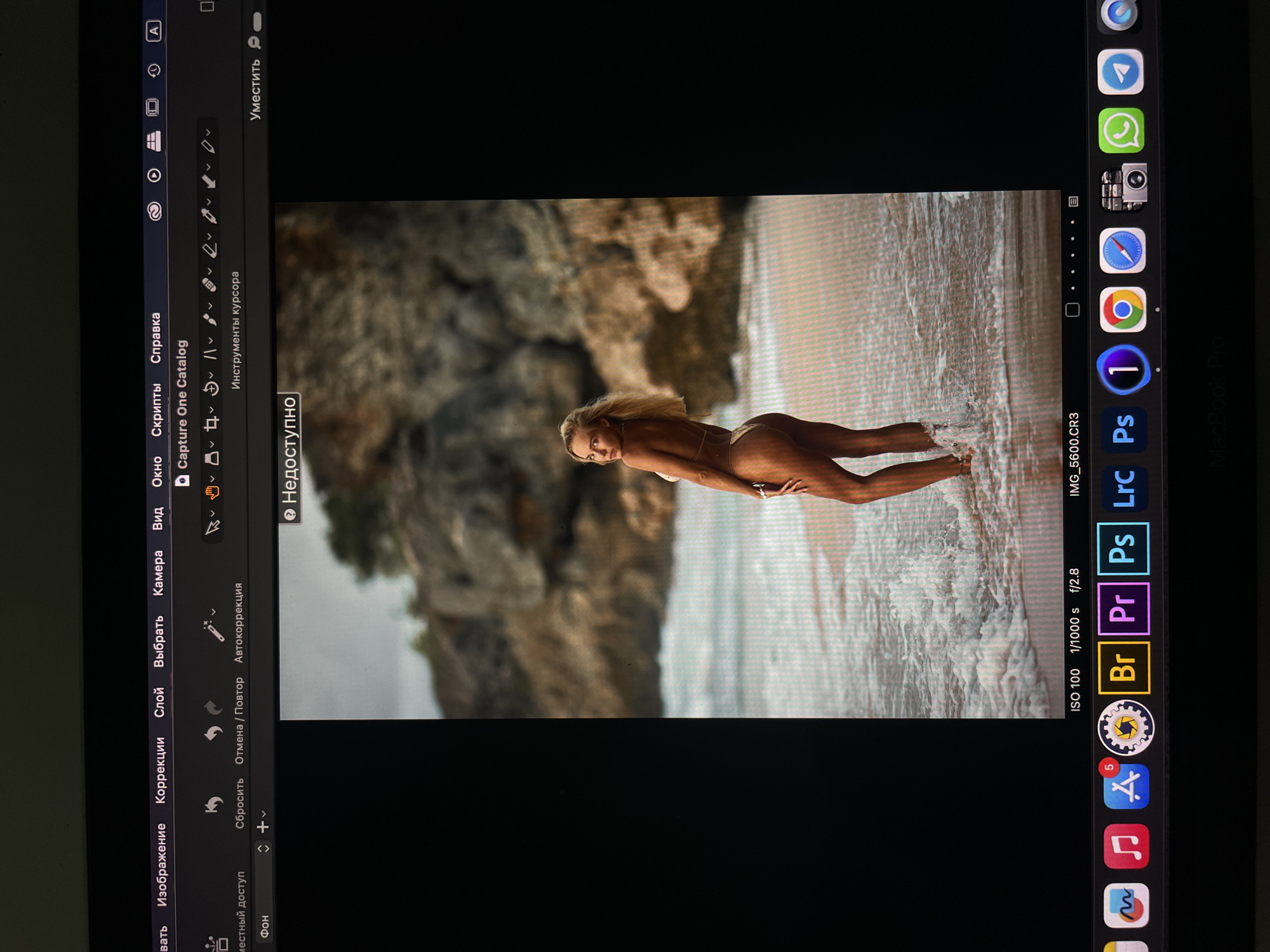 A screen displays a photo of a blonde woman in a bikini walking in the surf at a beach with a rocky backdrop. The screen shows the photo being edited with Capture One software. The ISO is 100, the shutter speed is 1/1000s, and the aperture is f/2.8. Icons on the screen indicate other programs such as Adobe Photoshop and Premiere Pro. The device appears to be a tablet. The image appears to be on a table. There is no indication that this scene is in Kazan, Russia.
