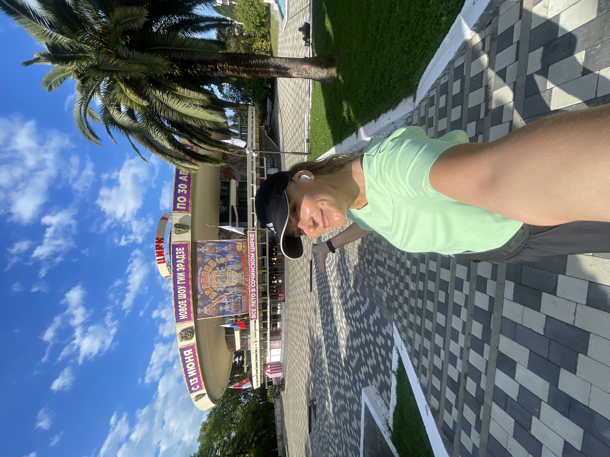 The image captures a woman taking a selfie in front of the Sochi State Circus in Sochi, Russia. She is wearing a light green t-shirt and a black baseball cap. The Sochi State Circus's facade, including a large banner advertising a show running from June 13 to August 30, is visible in the background. The sky is blue with some clouds. A palm tree stands to the left of the circus building. The ground is paved with a geometric pattern of grey and white blocks.
