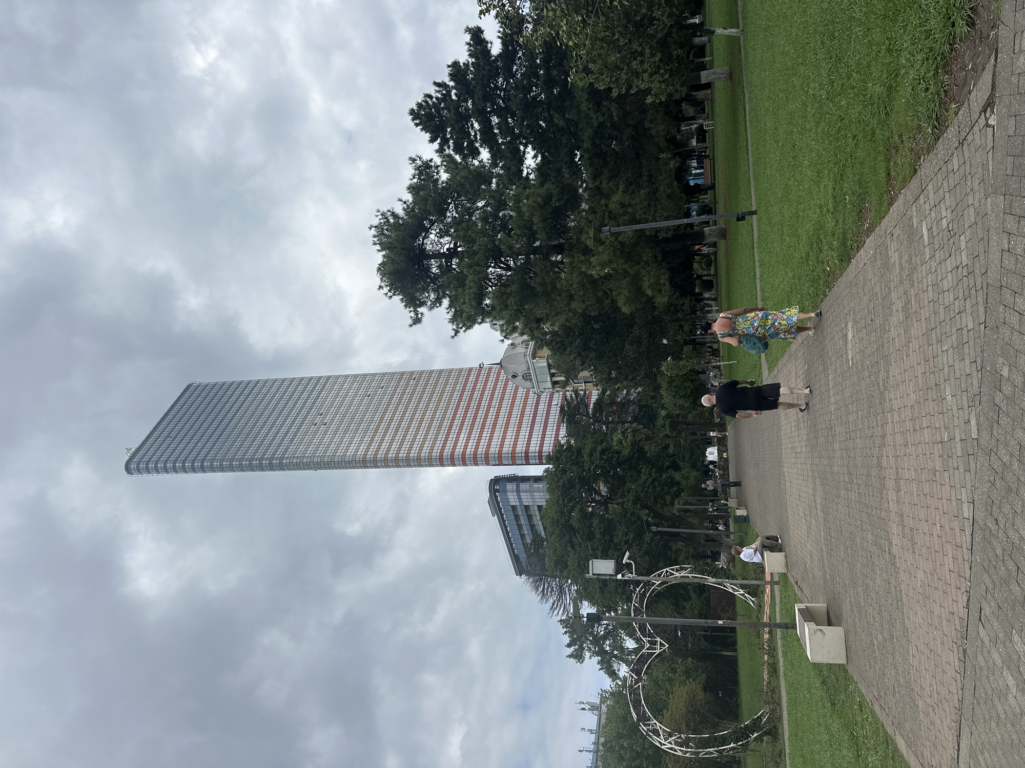 An overcast day in Batumi, Georgia, shows a wide, brick-paved walkway leading through a park. On the left, a large, illuminated heart-shaped sculpture is partially visible. Several benches are placed along the walkway. Two people are walking away from the camera, a man in black shorts and a t-shirt, and a woman in a floral dress carrying a blue bag. Further down the path, a woman in a white shirt and dark pants sits on a bench. In the background, tall trees form a dense green canopy. Beyond the trees, modern buildings dominate the skyline, most notably a very tall, slender skyscraper with a patterned facade of red and white horizontal stripes and glass. Another, shorter glass building with a curved design is situated to its left. The sky is filled with heavy grey clouds.