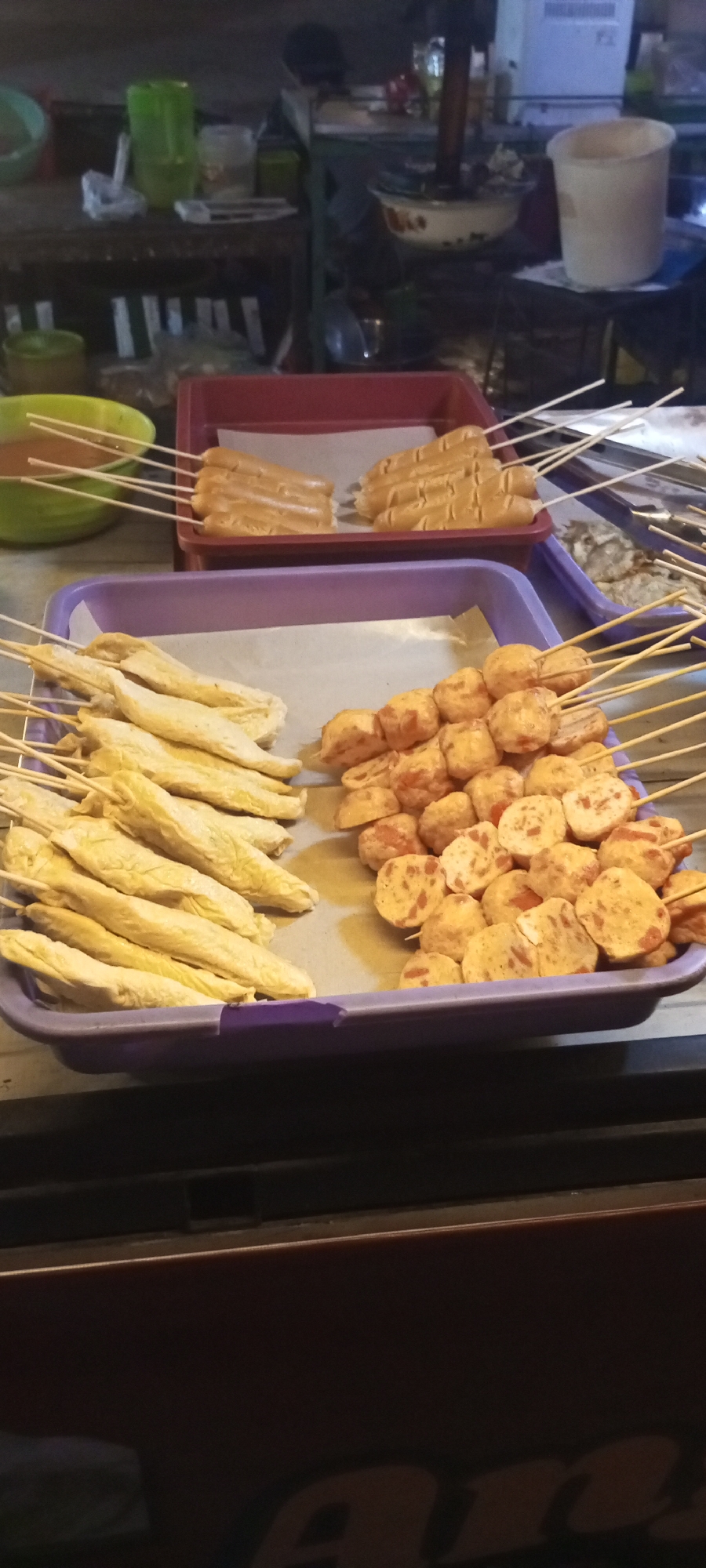A street food stall in Setu, Indonesia, displays two trays of Indonesian street food skewers. The tray in the foreground, on the right, holds skewers of round, fried fish cakes or meatballs, some sliced to reveal a reddish interior. To the left of this tray are skewers of elongated, pale yellow fish cakes or processed fish products. In the background, a red tray contains rows of brown, cylindrical sausages on skewers. A green bowl filled with a reddish sauce is partially visible on the left side of the image, with several skewers of food submerged in it. The stall appears to be set up outdoors, possibly at night, with dim lighting.