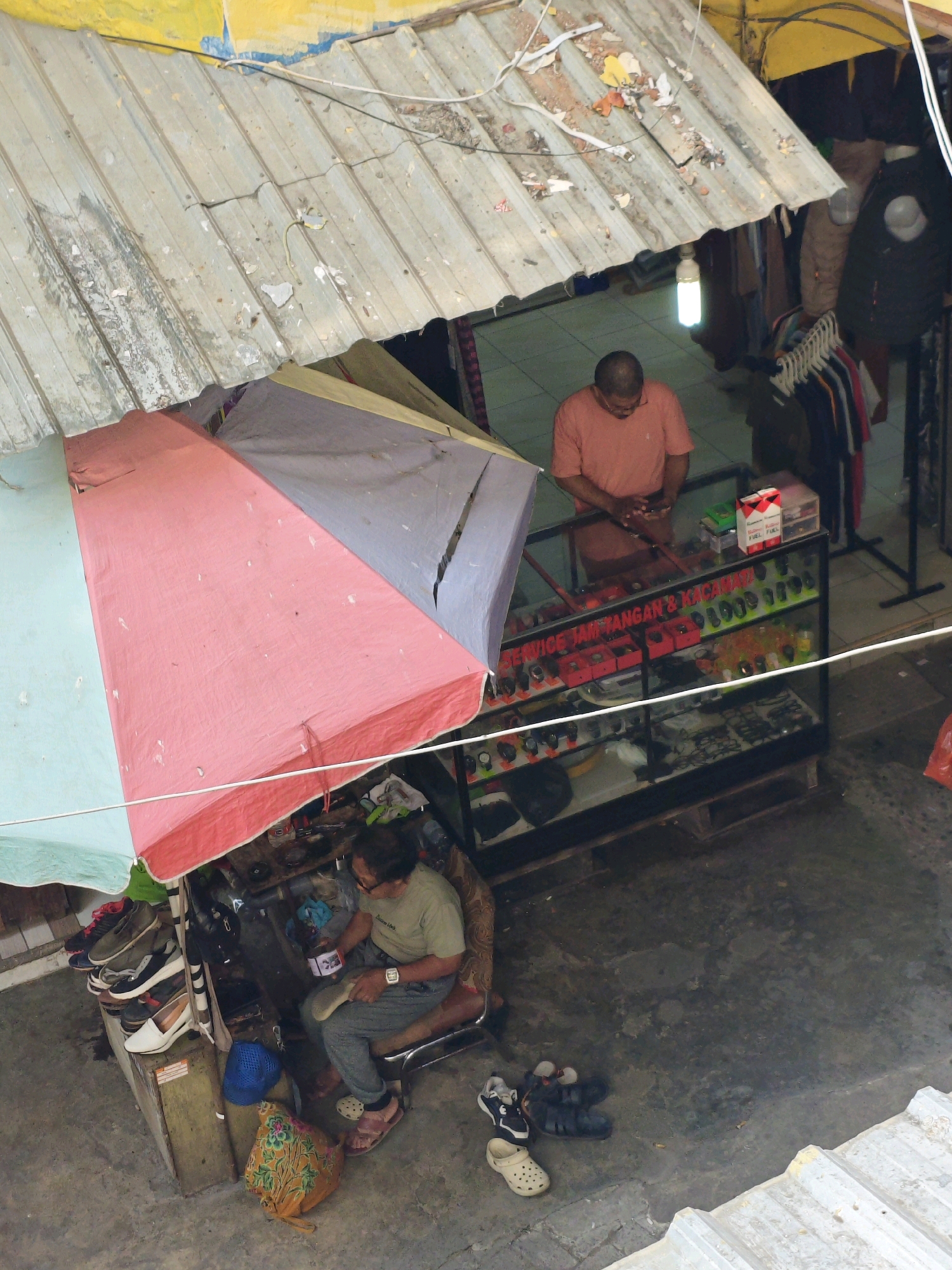 In Teddaopu, Indonesia, a shoe repair stall is depicted from an overhead perspective. The stall is covered by a weathered, corrugated metal roof supported by a wooden structure. A pink and light blue tarp is draped over the front, creating shade. Inside the stall, a man with glasses, wearing a light green t-shirt and grey pants, is seated on a stool. He is focused on repairing a shoe in his lap, holding a tool in his right hand. In front of him, there is a cluttered workspace with various shoe repair tools and supplies. To his right, stacked shoes are visible.
To the left of the shoe repair area, a glass display case is set up, containing watches and accessories. A man in an orange t-shirt stands behind the display case, looking down at his hands, which are holding a small object. The display case has a sign with red text that reads "SERVICE JAM TANGAN & KACAMATA". Several rows of watches are neatly arranged in the case, along with other items.
The ground around the stall is concrete. Scattered on the ground are various pairs of shoes, including sandals and sneakers. In the background, there are racks of clothing hanging, suggesting other goods are sold or displayed in the vicinity. The overall scene presents a humble, open-air marketplace setting.