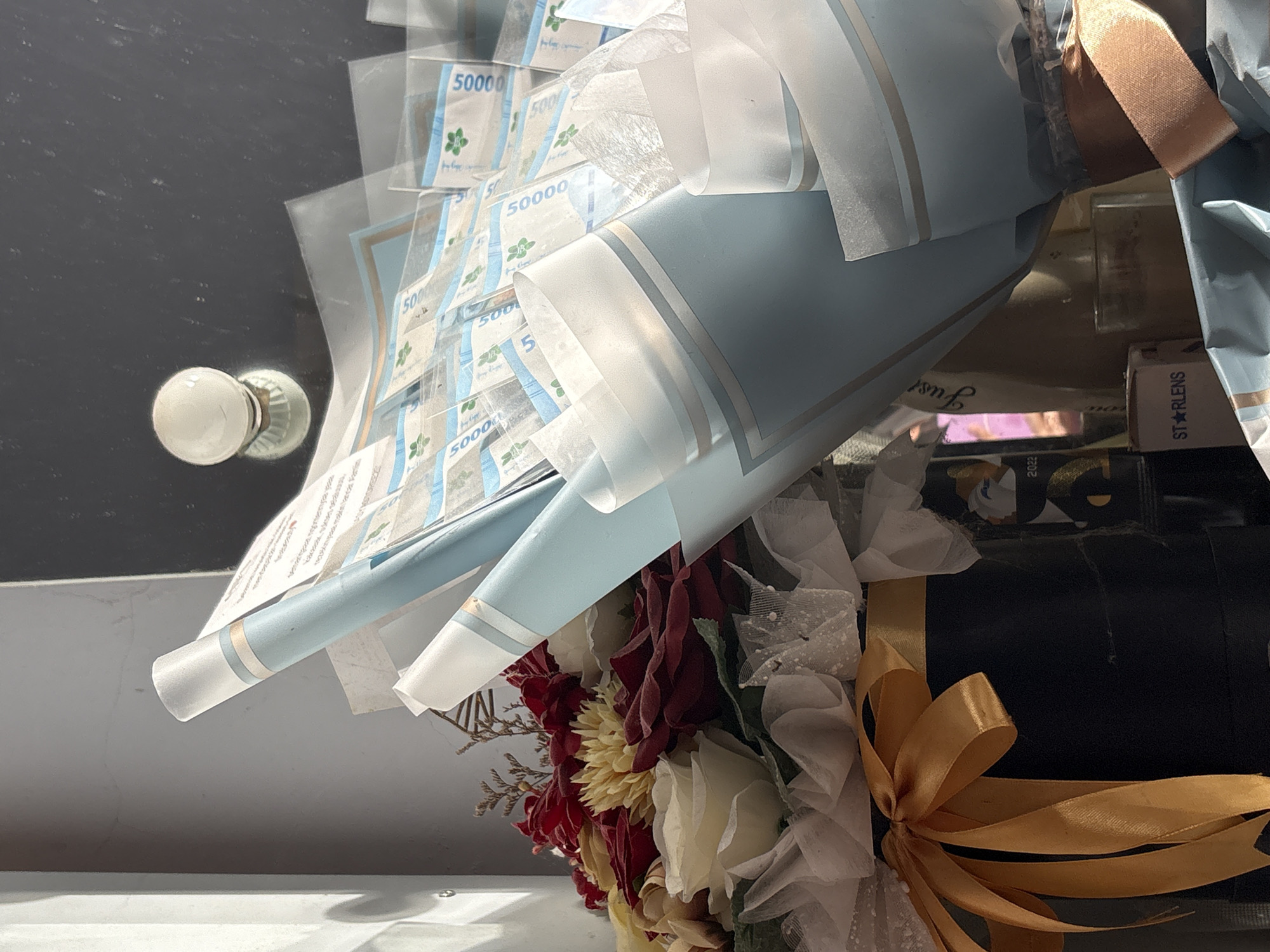 The image shows a floral and monetary bouquet. The bouquet contains flowers in shades of red, yellow, and white, interspersed with white netting and foliage. Wrapped around the flowers are what appear to be Indonesian Rupiah currency, specifically 50,000 IDR notes, encased in clear plastic sleeves and arranged like petals. The bouquet is wrapped in light blue paper with white stripes and tied with a gold ribbon. The background shows a light gray surface and an object, perhaps a wall with a light fixture mounted on it. A box is visible on the right with the text "STARLENS". Information about the location being Gadingrejo, Indonesia, is not directly discernible from the image but provided as additional context.