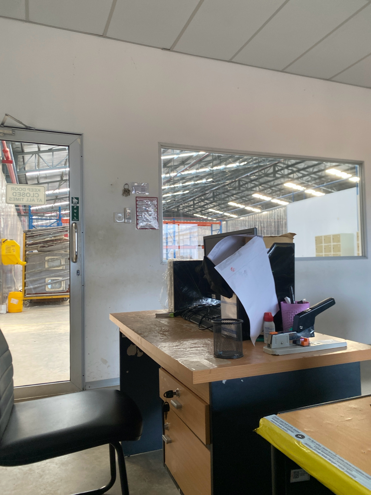 The image shows an office area within a warehouse in Batam, Indonesia. A desk with a computer monitor, stapler, and other office supplies is positioned in the foreground, next to a black office chair. Through a large window, the interior of the warehouse is visible, filled with rows of shelving units and stacked materials. A glass-paneled door with a "PULL" sign is to the left, leading to the warehouse floor. The walls are painted white, and the ceiling is composed of acoustic tiles. The overall impression is of a functional workspace within an industrial environment.