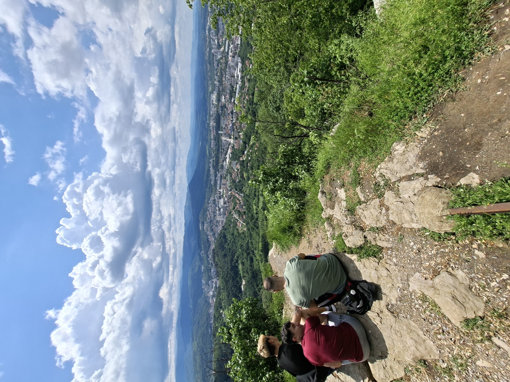 The image depicts a high-angle view of three individuals seated on a rocky outcrop overlooking the city of Gabrovo, Bulgaria. The sky is visible, featuring white, puffy clouds against a blue backdrop. The urban area below consists of buildings, streets, and green spaces stretching into the distance. The foreground showcases the edge of the rocky terrain covered with grass and plants. Three people are visible: one wearing a green shirt, another in a red shirt, and the third person is wearing a black shirt. They are sitting and appear to be taking in the view. A bag is on the ground near the individual in the green shirt.