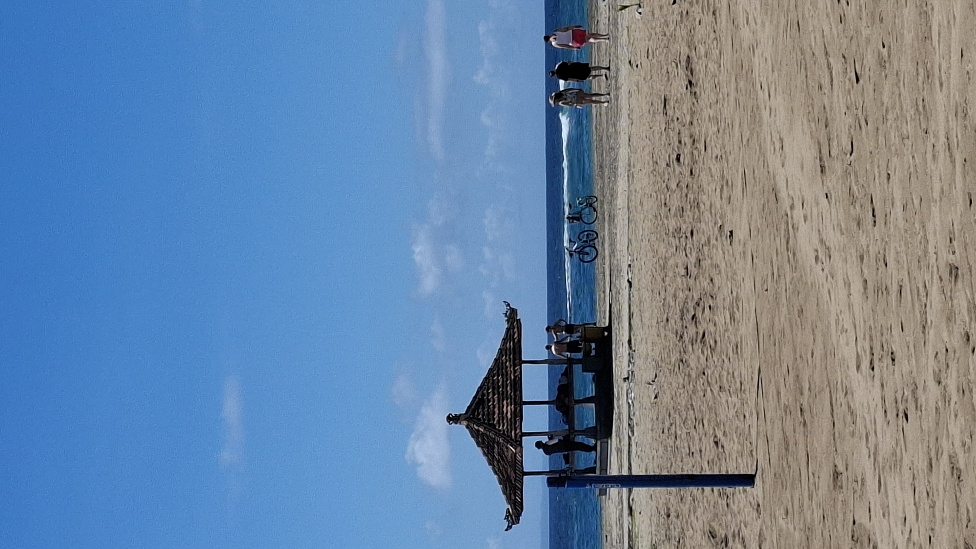 The image depicts a beach scene in Kuta Selatan, Indonesia. A thatched-roof structure stands on the sand, with several people seated inside. Further down the beach, near the water's edge, are a couple of bicycles and several people walking. The sky is blue with a few scattered clouds. The water is visible, creating a horizontal line. The sand is a light tan color, and the texture of the wood to the right suggests a wooden boardwalk. A dark blue metal bar is partially visible, vertically placed.